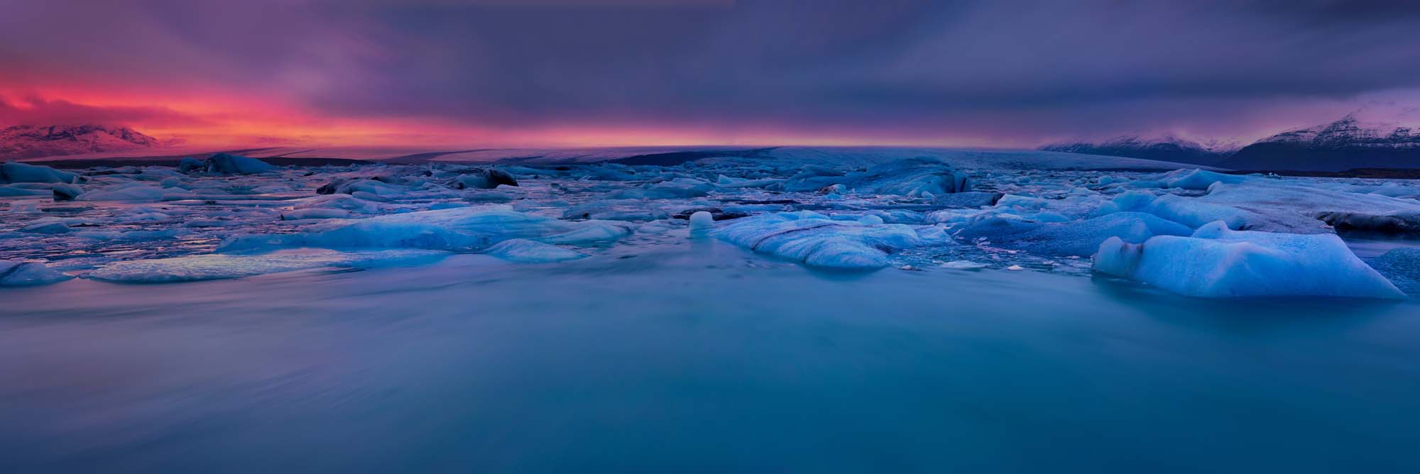 A stunning twilight panorama of Iceland's Jokulsarlon glacier lagoon, featuring icebergs floating in crystalline turquoise water beneath a dramatic sunset sky painted in shades of crimson, purple, and deep blue. The surreal landscape captures the raw beauty of Iceland's glacial region during the magical golden hour.