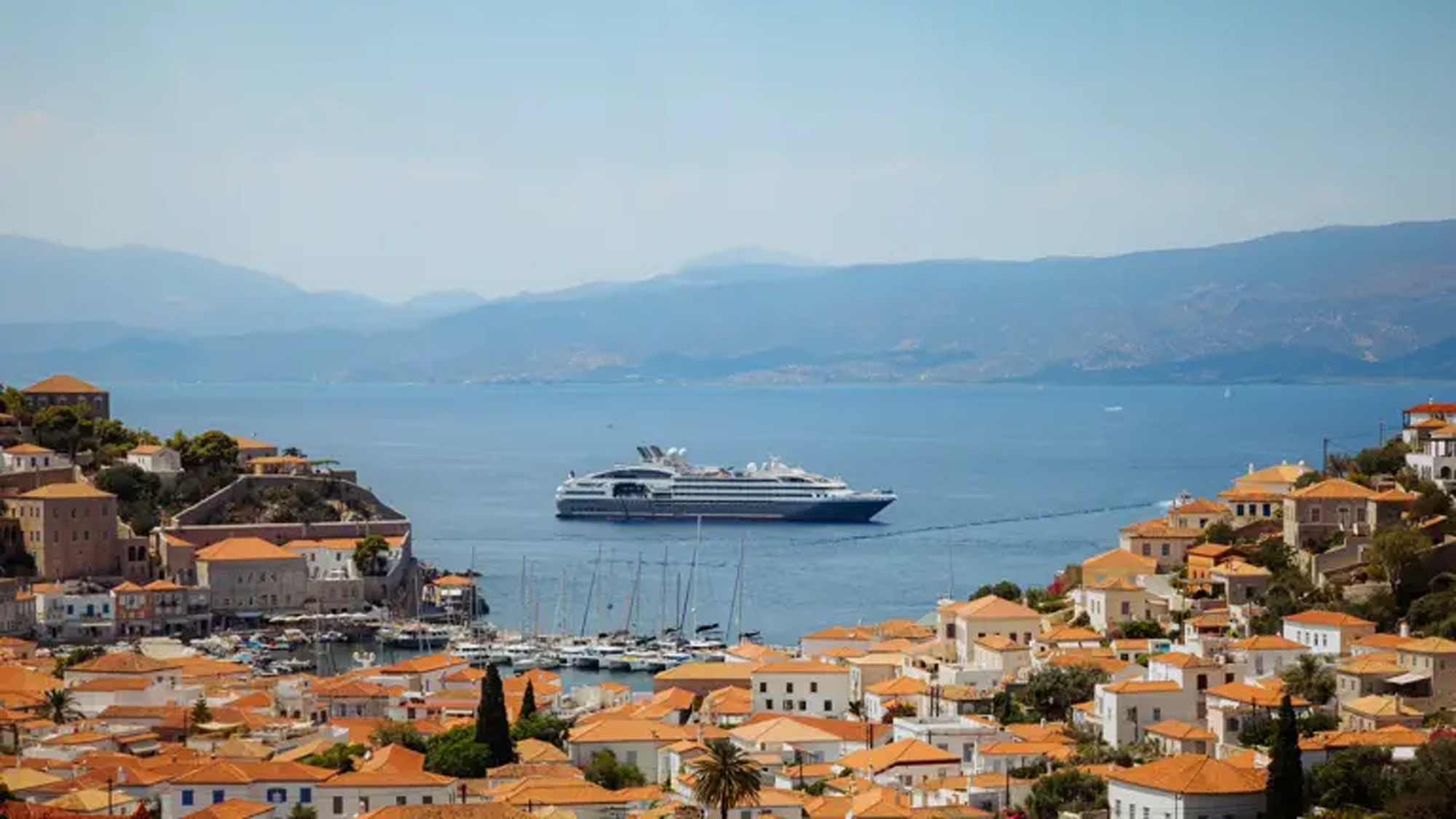 A large modern cruise ship anchored in the crystalline Mediterranean waters off the picturesque Greek island of Hydra, with traditional whitewashed buildings featuring orange-tiled roofs cascading down the hillside, sailboats moored in the harbor, and the mainland mountains visible across the water in the distance.