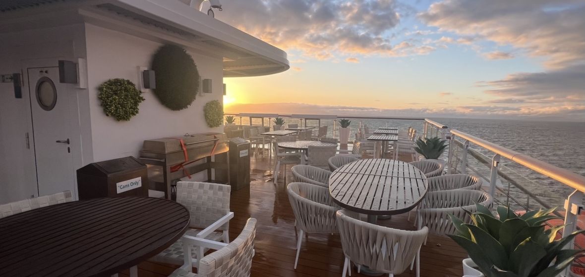 Cruise Ship Lido Deck at Sunset An expansive outdoor lido deck aboard a cruise ship featuring multiple dining tables with white woven chairs, decorative spherical planters, and a commanding view of the ocean at golden hour. The deck extends toward the horizon with calm seas and a dramatic sunset sky, showcasing the ship's upper deck amenities for dining and relaxation.