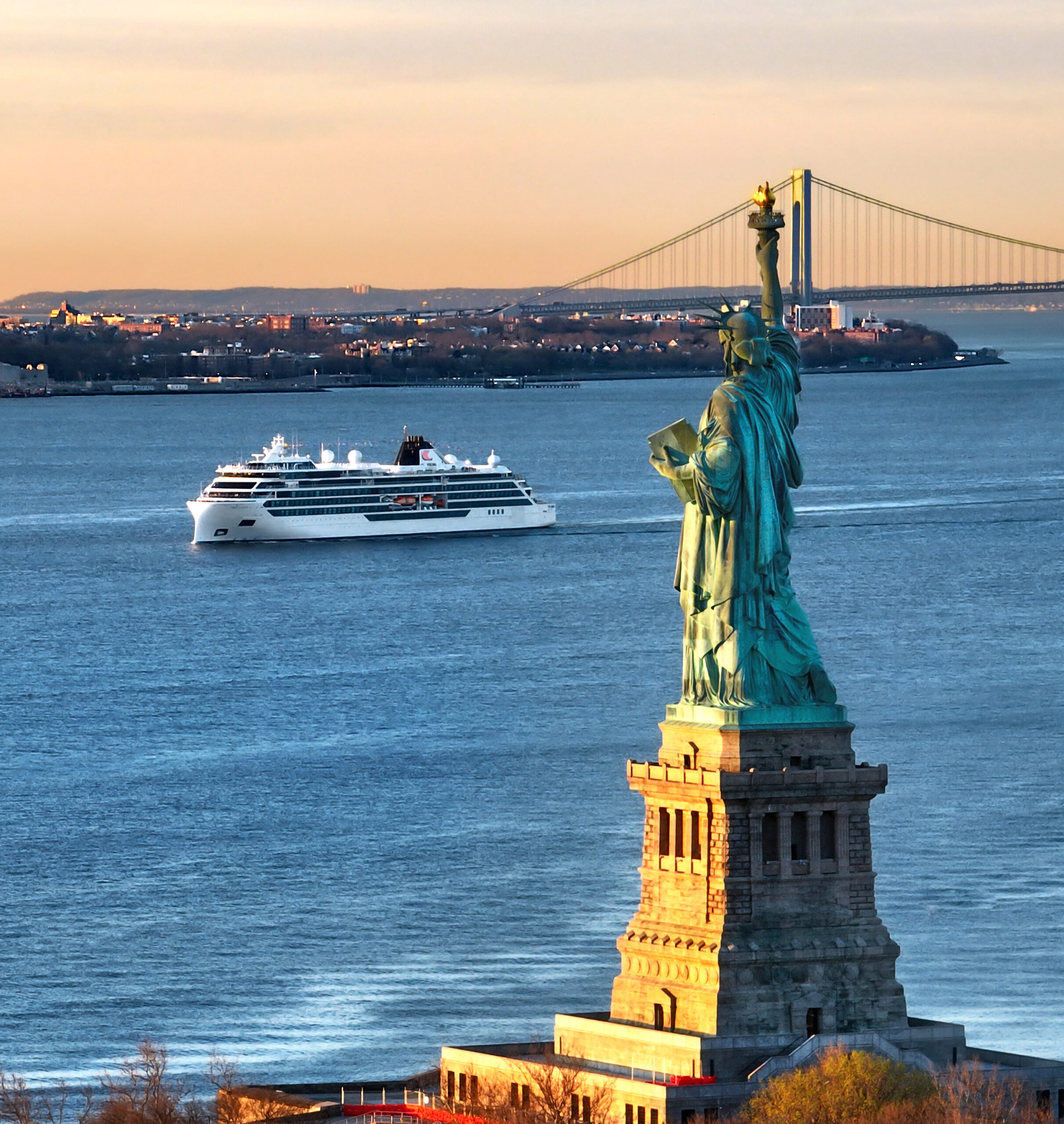 A modern cruise ship enters New York Harbor past the iconic Statue of Liberty on Liberty Island, with the Verrazano-Narrows Bridge visible in the background and the Manhattan skyline dotted with buildings along the shoreline. The scene captures a golden-hour moment with warm sunlight illuminating the statue's copper patina and the ship's white hull against the blue harbor waters.