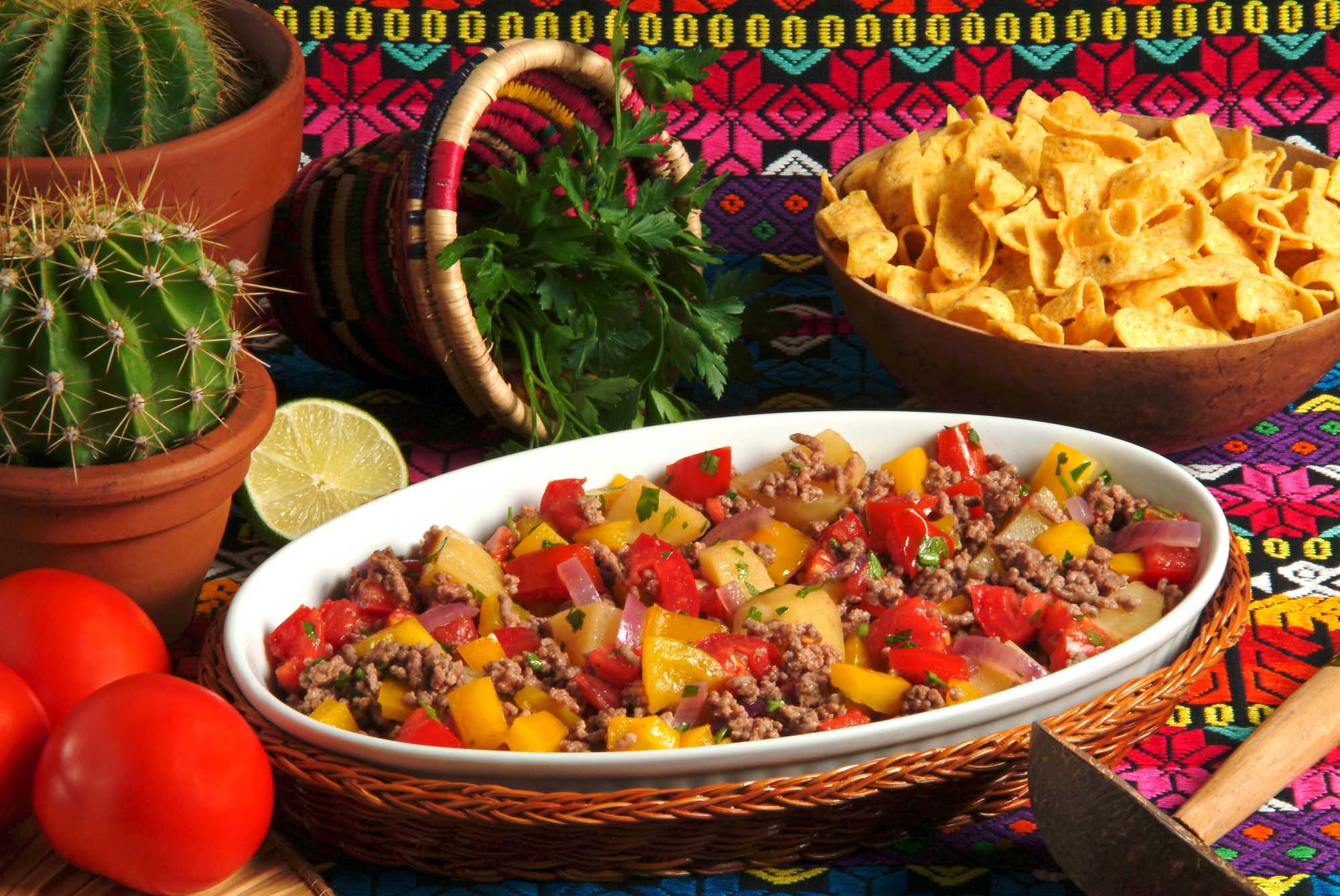 A vibrant presentation of Cuban picadillo habanero served in a white ceramic bowl, featuring seasoned ground meat with diced potatoes, red and yellow peppers, and fresh cilantro garnish. The dish is artfully arranged on a colorful woven placemat alongside complementary items including fresh lime, a decorative ceramic pitcher, potted cactus, crispy tortilla chips, and fresh tomatoes, creating an authentic and appetizing Mexican-Cuban culinary scene.