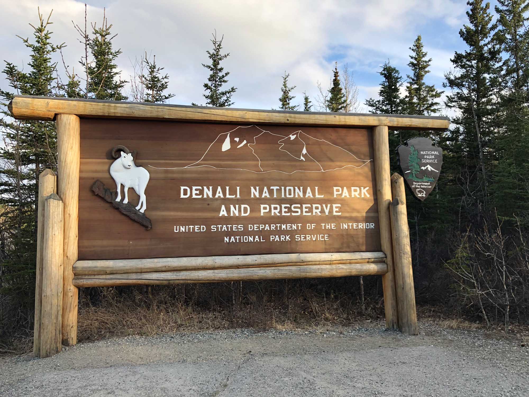 A rustic wooden entrance sign for Denali National Park and Preserve in Alaska, featuring a white Dall sheep illustration and mountain outline artwork. The sign is crafted from natural logs and displays official National Park Service branding, surrounded by boreal forest vegetation.