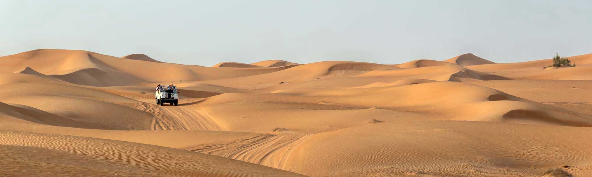 A white SUV navigates through rolling golden sand dunes in a vast desert landscape, with the vehicle creating distinctive tire tracks across the rippled sand. The dramatic lighting emphasizes the undulating topography and the solitary nature of desert exploration.
