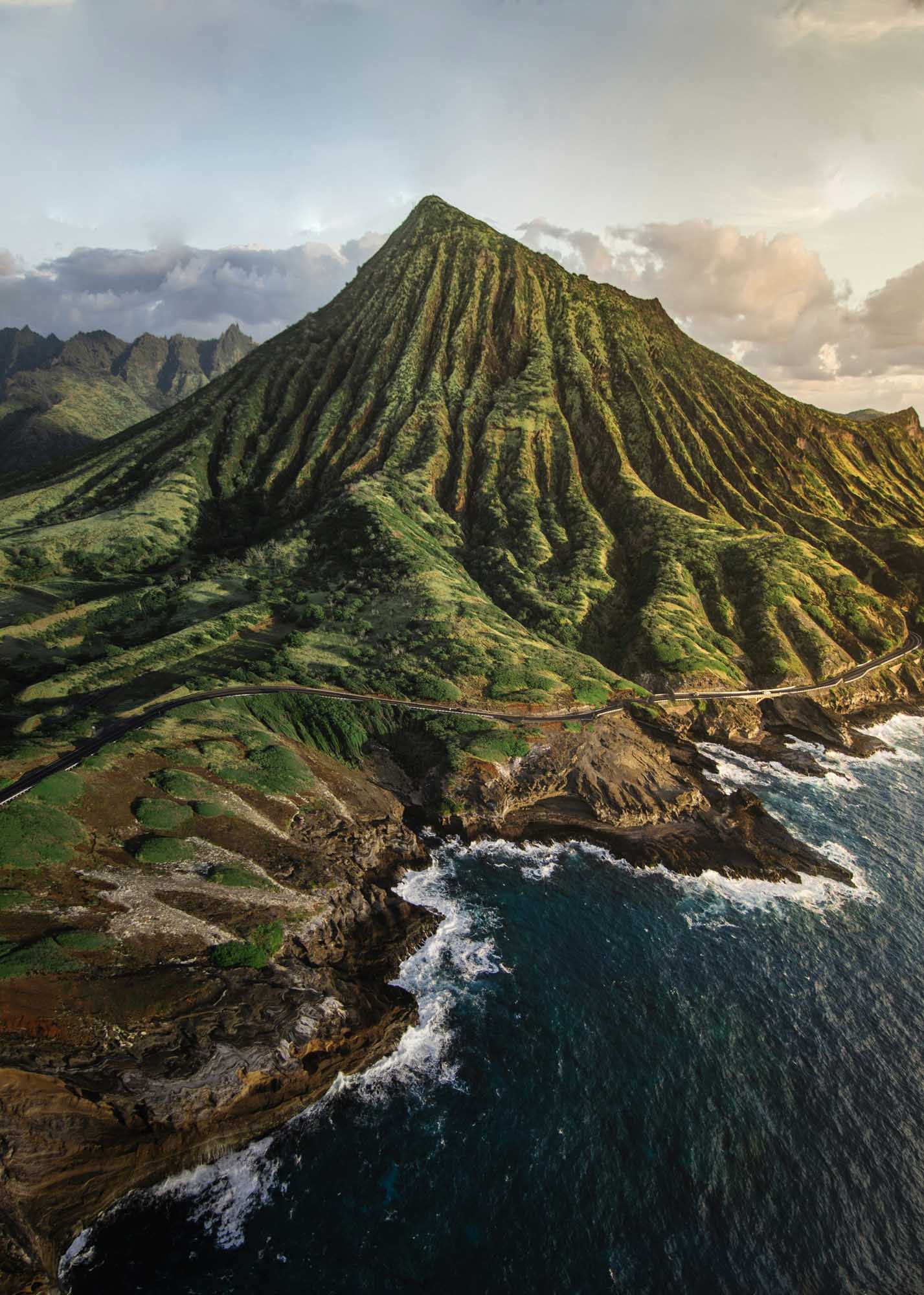 Dramatic Coastal Mountain with Fluted Ridges A stunning aerial view of a dramatically fluted volcanic mountain rising from the Hawaiian coastline, with distinctive erosion patterns creating deep vertical ridges across its lush green slopes. Waves crash against the rugged cliffs below while other verdant peaks frame the landscape in the background.
