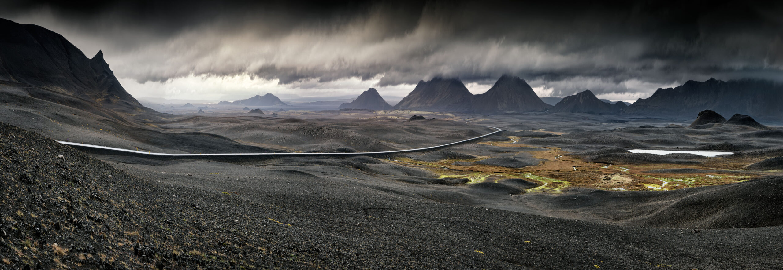 A sweeping panoramic view of Iceland's stark volcanic highlands, featuring a winding glacial river cutting through dark lava fields, dramatic jagged mountain peaks in the distance, and moody storm clouds overhead. The landscape showcases the raw, otherworldly terrain characteristic of Iceland's interior, with patches of golden vegetation dotting the desolate terrain.