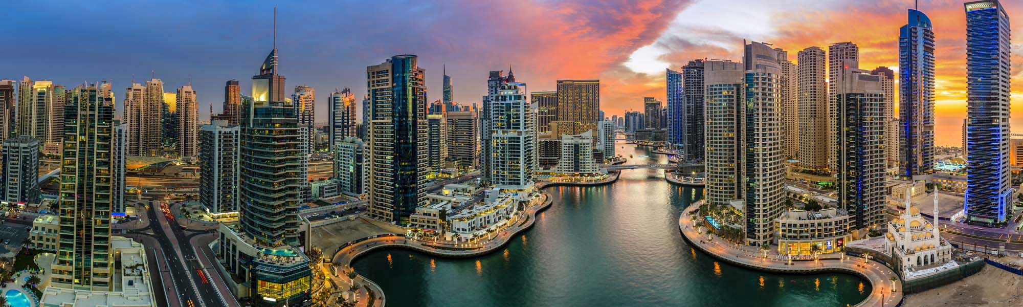 A stunning panoramic view of Dubai Marina at golden hour, showcasing the iconic waterfront development with modern high-rise residential and commercial towers reflected in the calm marina waters. The vibrant sunset sky in shades of orange and gold illuminates the contemporary architecture and bustling waterfront promenade lined with restaurants and entertainment venues.