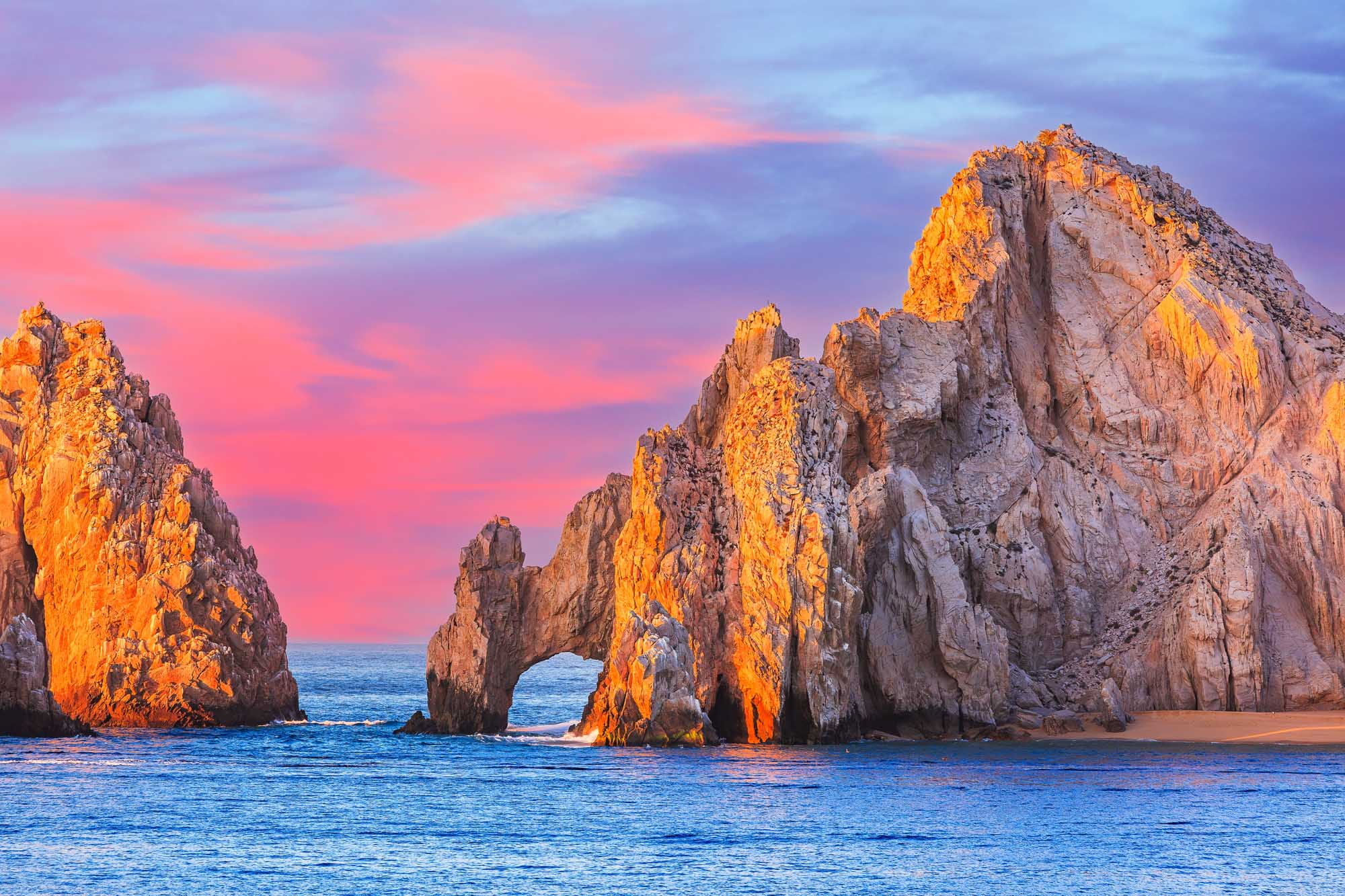 The iconic El Arco (The Arch) rock formation at Cabo San Lucas is bathed in golden sunset light, with dramatic granite cliffs rising from the turquoise Pacific waters. The striking natural landmark features a natural arch carved through the rock, creating a spectacular gateway between the land and sea, with vibrant pink and purple hues filling the sky.