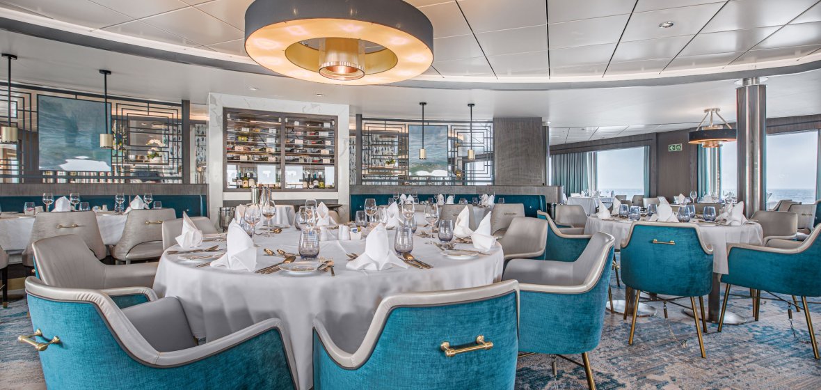 Elegant Fine Dining Restaurant with Turquoise Accents A sophisticated cruise ship dining venue featuring round tables dressed in white linens, modern turquoise and gray upholstered chairs with gold accents, and a contemporary interior with geometric wall patterns, ambient lighting, and floor-to-ceiling windows. The refined setting showcases premium furnishings and elegant table settings characteristic of luxury cruise accommodations.