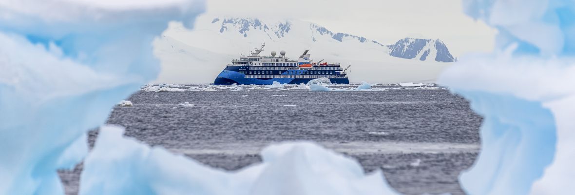 Expedition Cruise Ship in Antarctic Waters A modern expedition cruise ship navigates through icy Antarctic waters, framed by massive icebergs and snow-covered peaks in the background. The vessel features distinctive blue hull livery and is positioned among pristine polar ice formations under a partly cloudy sky.