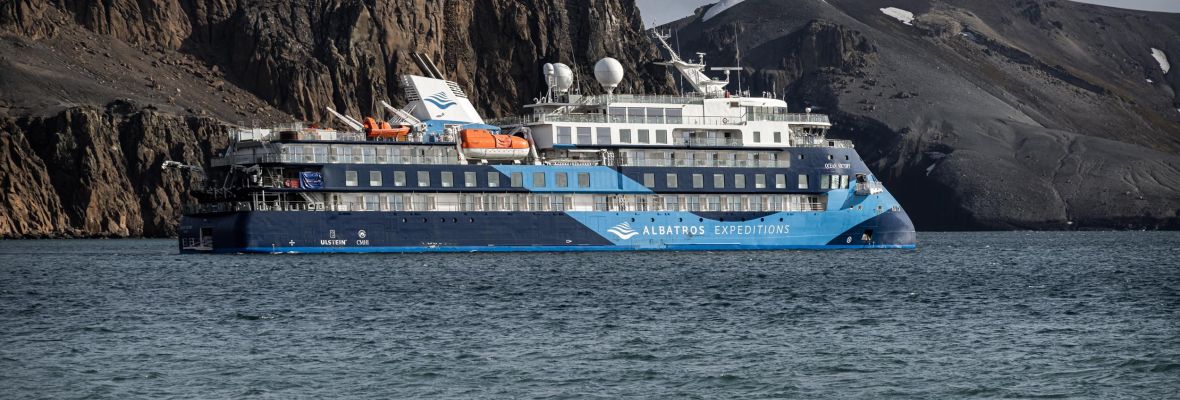 Expedition Ship at Deception Island, Antarctica An expedition cruise ship with distinctive blue and white livery is anchored in the waters off Deception Island, Antarctica, with dramatic volcanic cliffs and rocky terrain visible in the background. The vessel appears to be a purpose-built polar expedition ship designed for remote destination cruising in extreme environments.