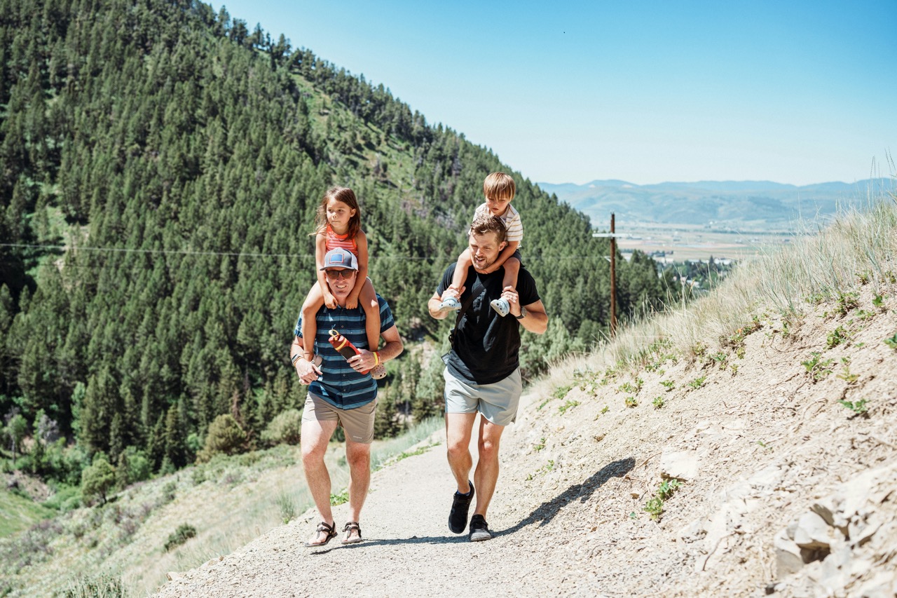 A father carrying his young son on his shoulders runs down a scenic dirt trail through a dense coniferous forest, with two children jogging alongside him. The expansive valley and distant mountains are visible in the background under clear blue skies, capturing an active outdoor family adventure in a mountainous region.