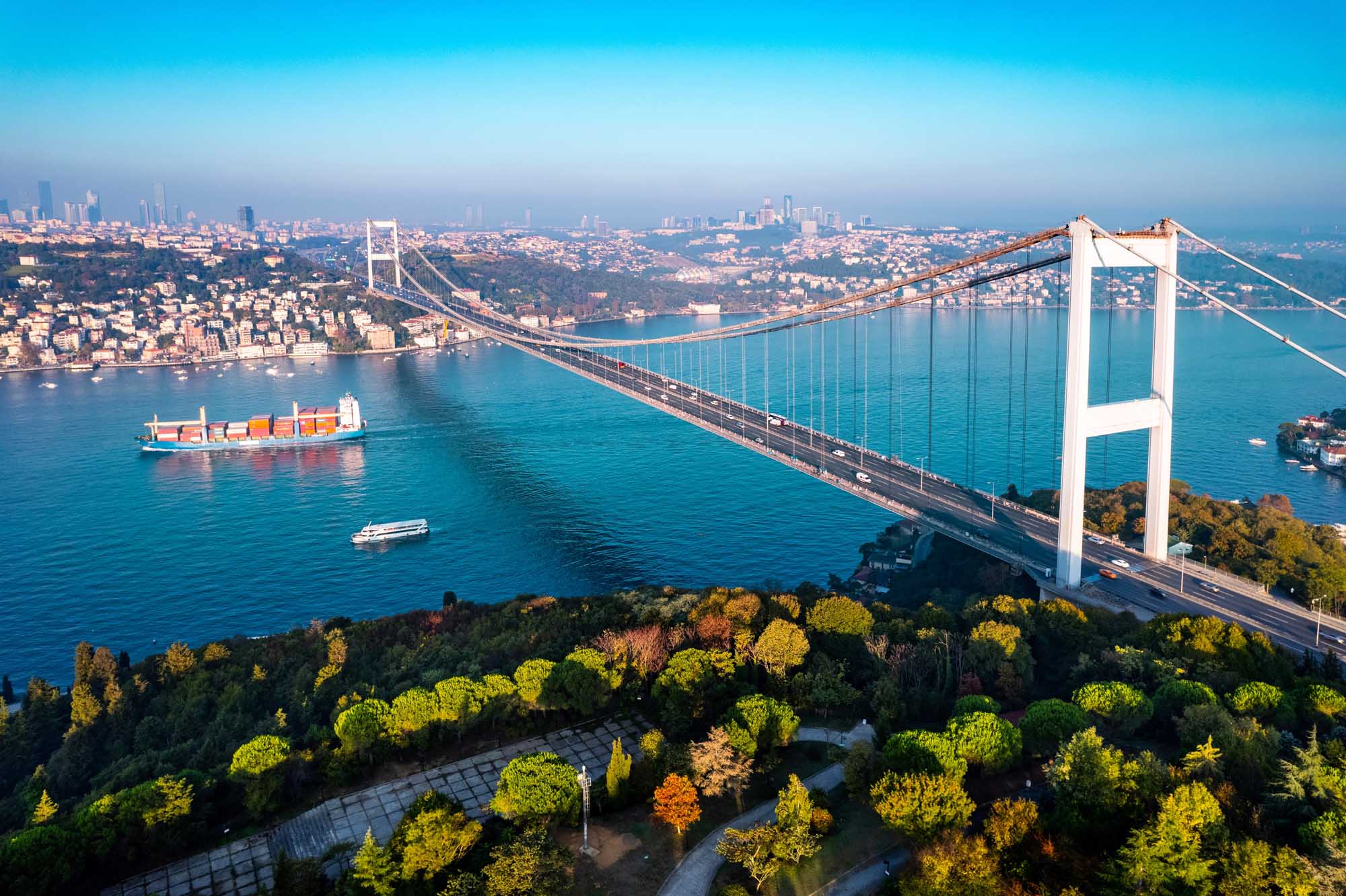 Fatih Sultan Mehmet Bridge Over Istanbul Bosphorus An aerial view of the iconic Fatih Sultan Mehmet Bridge spanning the Bosphorus Strait in Istanbul, Turkey, with a cargo ship and ferry navigating the turquoise waters below, the Asian coastline visible in the distance, and autumn-colored forests in the foreground.