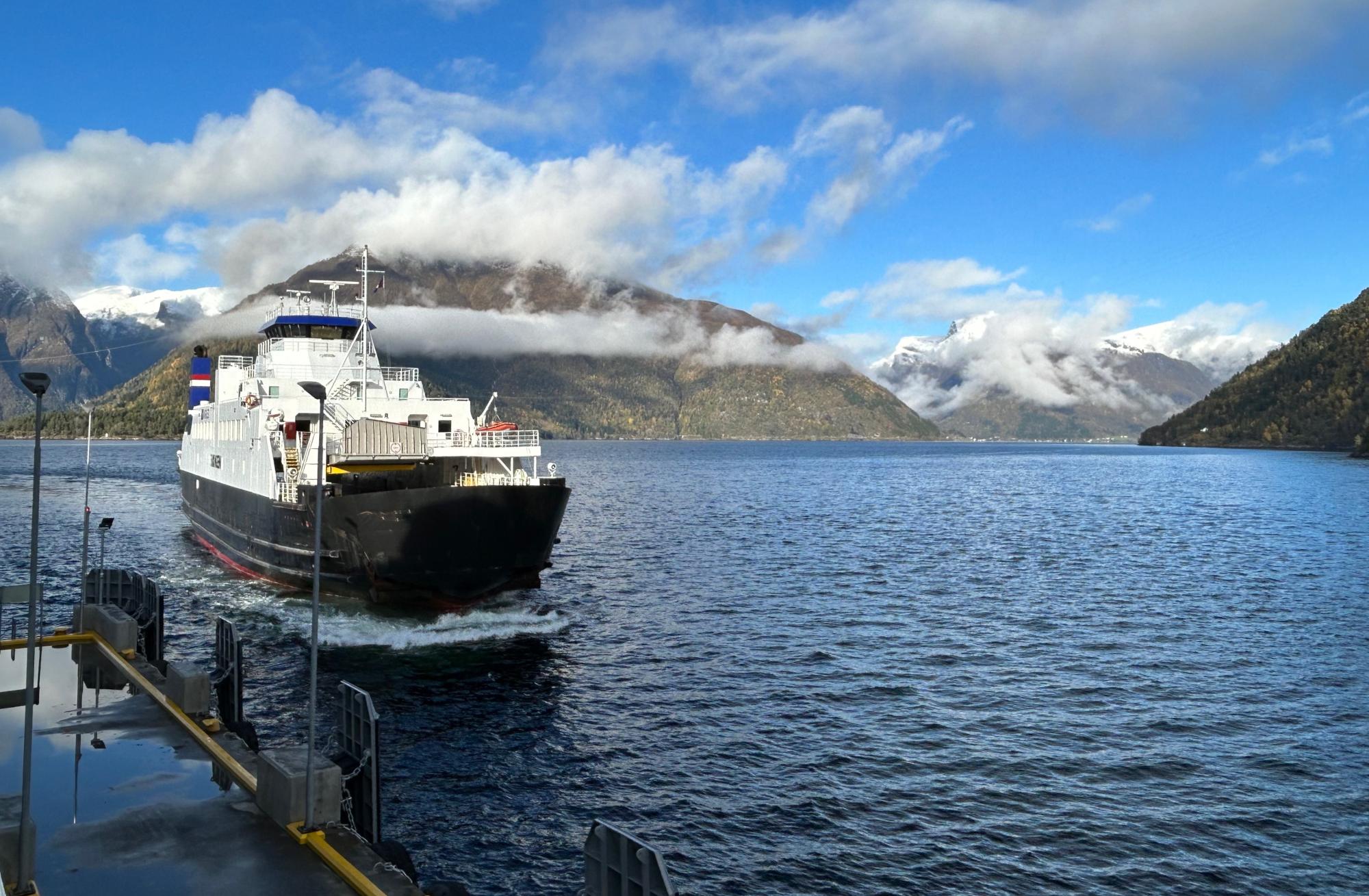 Ferry Departing Patagonian Lake with Snow-Capped Mountains A white and black passenger ferry with Dutch flag markings departs from a dock on a pristine alpine lake, surrounded by dramatic snow-covered mountains partially shrouded in mist under a bright blue sky. The scenic landscape features steep forested slopes and glaciated peaks characteristic of Patagonia.