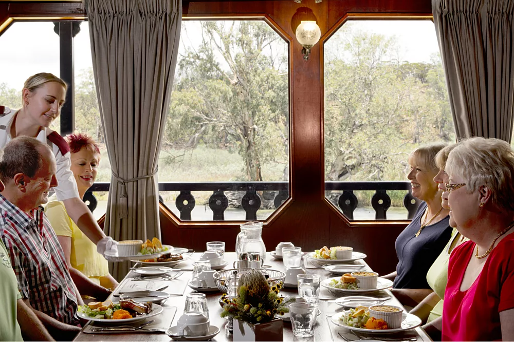 An elegant formal dining room aboard a cruise ship with passengers enjoying a meal at a long table. Large windows frame scenic forest and landscape views, while gray curtains, wooden paneling, and warm lighting create a sophisticated atmosphere.