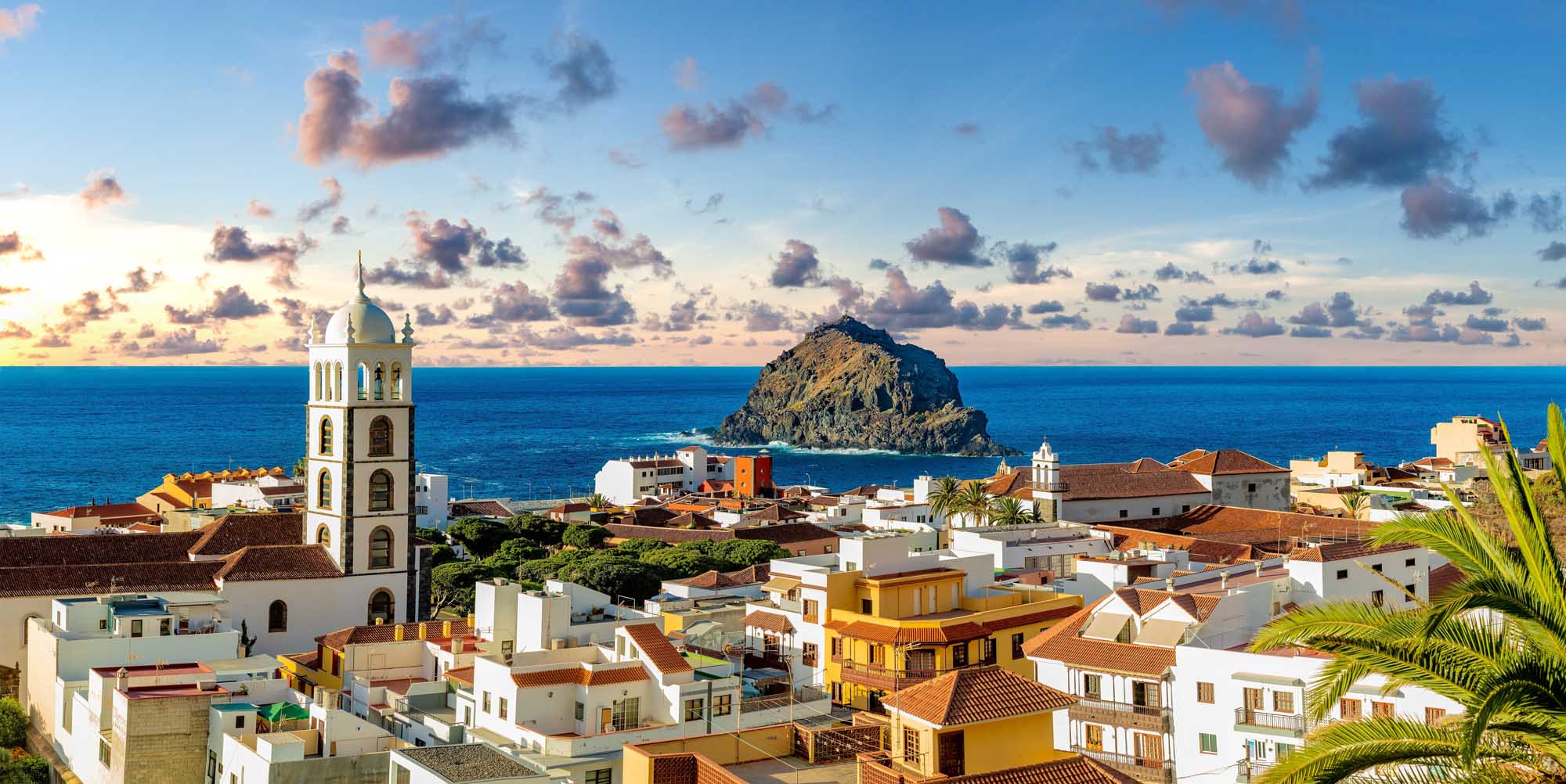 A picturesque panoramic view of the historic coastal village of Garachico in Tenerife, Canary Islands, featuring colorful whitewashed and yellow buildings with terra cotta roofs, a prominent church bell tower, and a distinctive dark volcanic rock formation rising from the turquoise Atlantic Ocean under a dramatic cloud-filled sky at sunset.