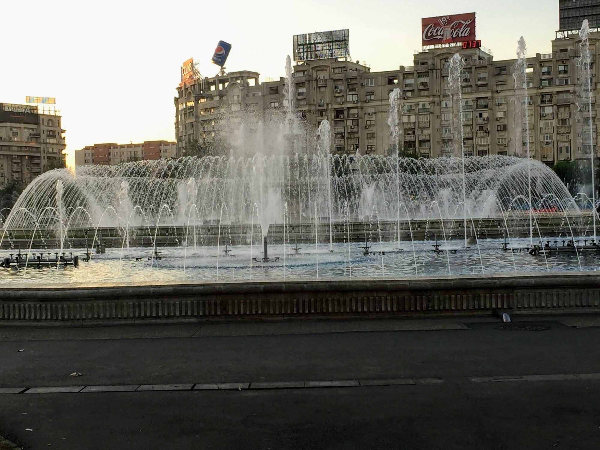 Grand Fountain Display in Soviet Square A spectacular large-scale fountain with multiple water jets and arching sprays in an urban plaza, featuring Soviet-era apartment buildings in the background and prominent vintage signage including a Coca-Cola advertisement on the roofline. The architectural style and urban design are characteristic of mid-20th century Eastern European cities.