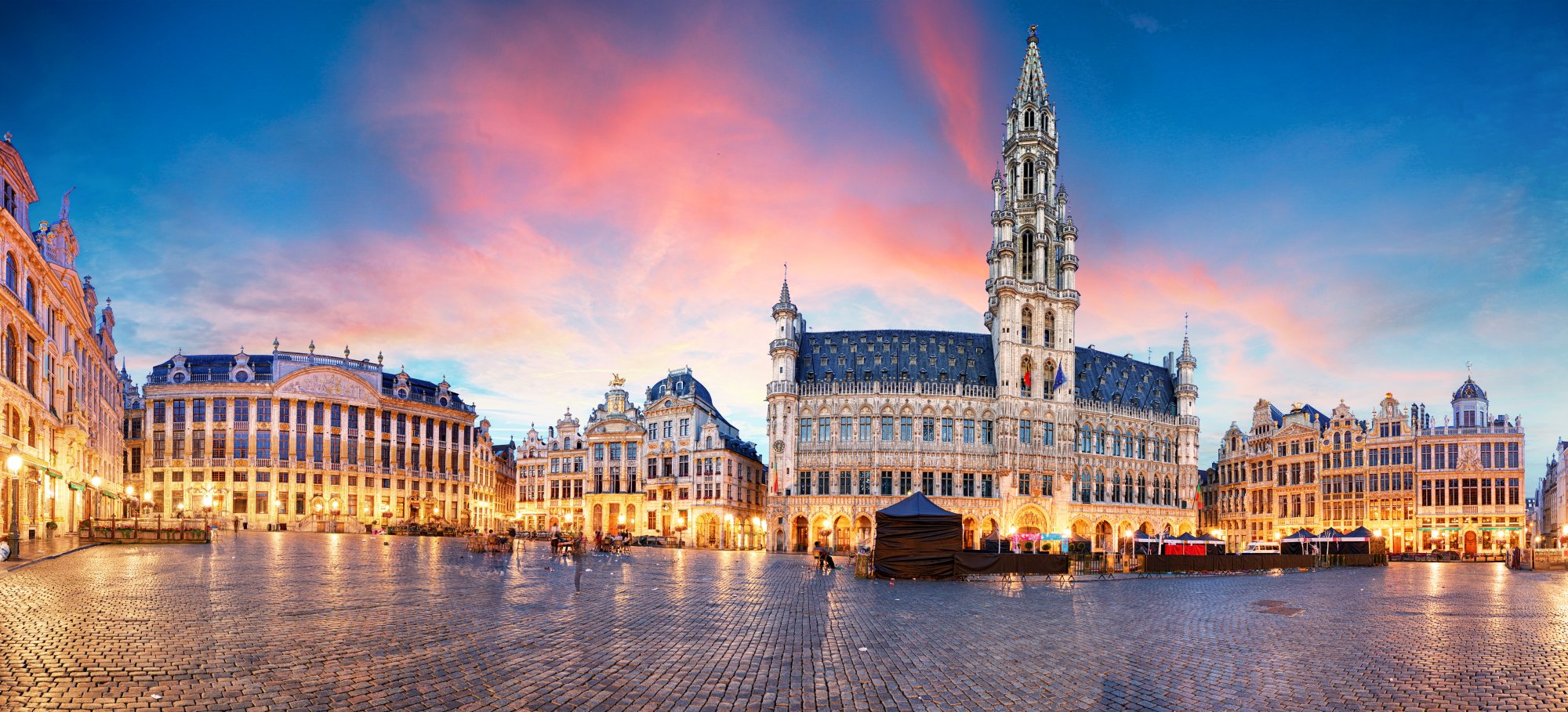 Grand Place Brussels at Twilight A stunning twilight view of Brussels' iconic Grand Place (Grote Markt) featuring the ornate Gothic Town Hall with its distinctive spire, surrounded by elegant 17th-century guild houses illuminated by warm golden lights against a vibrant blue and pink sunset sky. The cobblestone plaza is wet from rain, reflecting the architectural splendor of Belgium's most famous square.