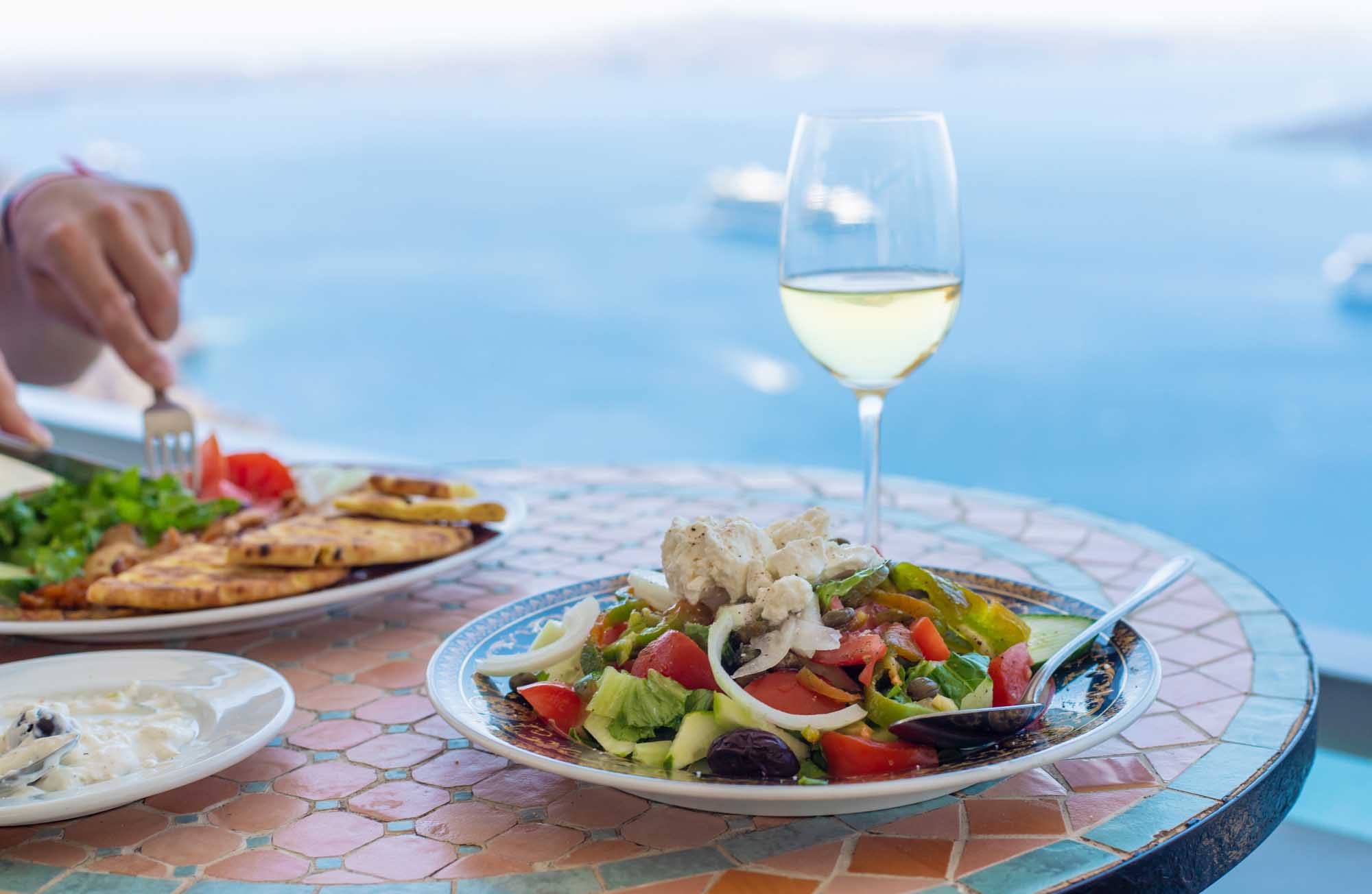 Greek Salad and White Wine Mediterranean Dining A scenic Mediterranean dining scene featuring a fresh Greek salad with feta cheese, olives, and vegetables on a decorative plate, paired with a glass of white wine. The table overlooks a stunning blue sea and sky, capturing the essence of Greek island hospitality and coastal leisure dining.