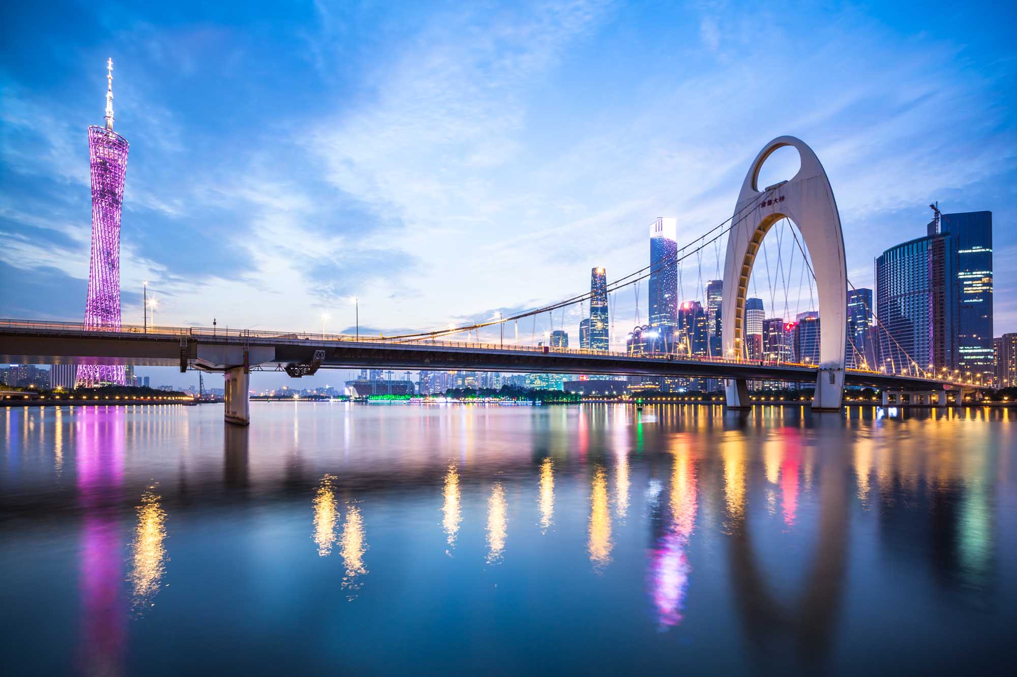 Guangzhou Skyline with Illuminated Bridges at Dusk A breathtaking twilight cityscape of Guangzhou, China, featuring the iconic Liede Bridge spanning across the Pearl River. The scene captures the city's modern skyline illuminated with golden and pink lights, with a distinctive pink-lit tower on the left and a prominent white arch bridge on the right, all beautifully reflected in the calm waters below.