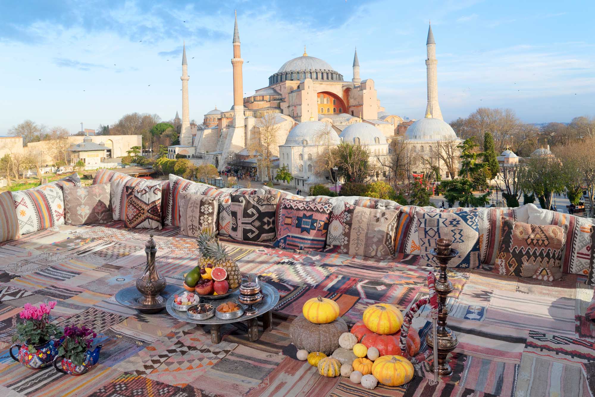 Hagia Sophia with Rooftop Lounge in Istanbul A stunning rooftop terrace featuring traditional Turkish cushions, kilim textiles, and autumn produce overlooking the iconic Hagia Sophia mosque in Istanbul, Turkey. The scene captures the architectural grandeur of the historic landmark with its massive dome and minarets against a clear blue sky.