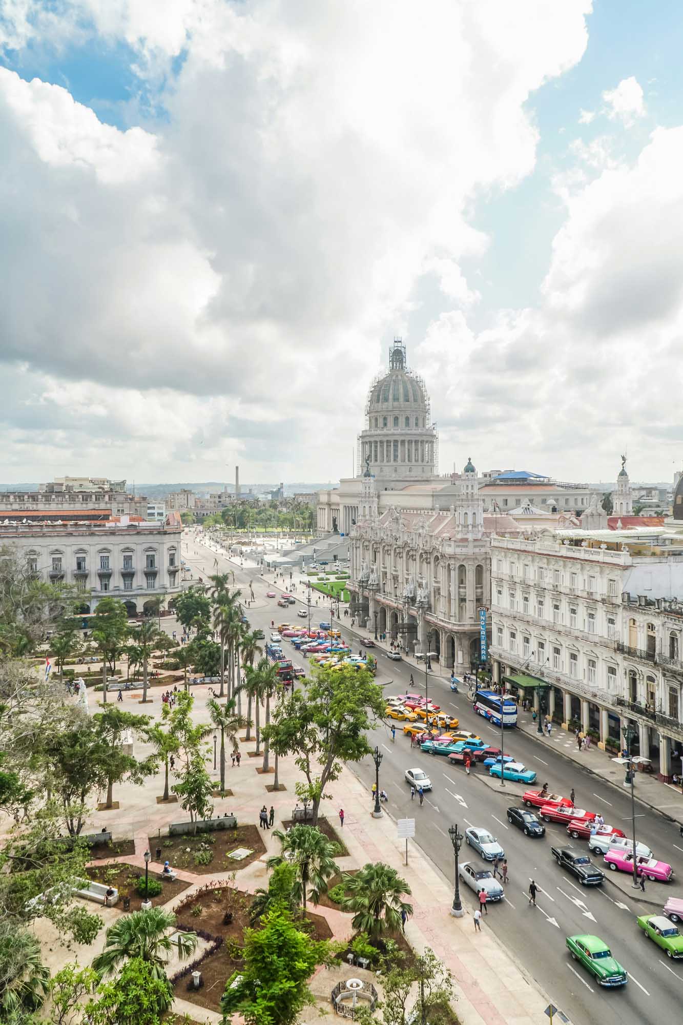 An elevated daytime view of Havana's iconic Capitol building dominating the skyline, with its distinctive dome prominently featured. The street below bustles with colorful classic American cars and palm-lined plazas, capturing the vibrant energy of Cuba's capital city.