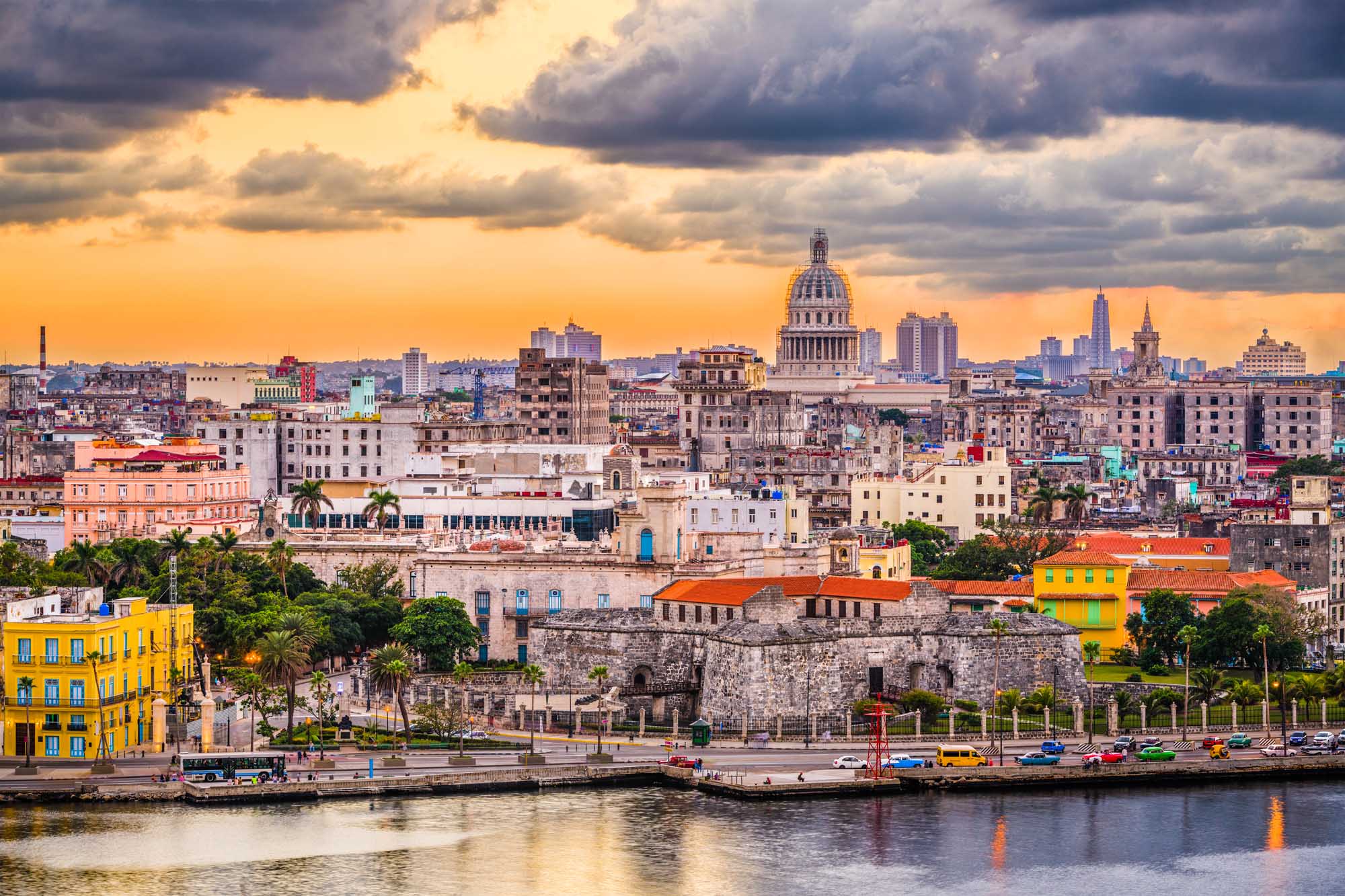A stunning panoramic view of Havana, Cuba's downtown skyline at dusk, featuring the iconic Capitol building with its distinctive dome, colorful colonial architecture, palm trees, and the waterfront promenade (Malecón) along the harbor. The dramatic golden sky with storm clouds creates a striking backdrop for this vibrant Caribbean city.