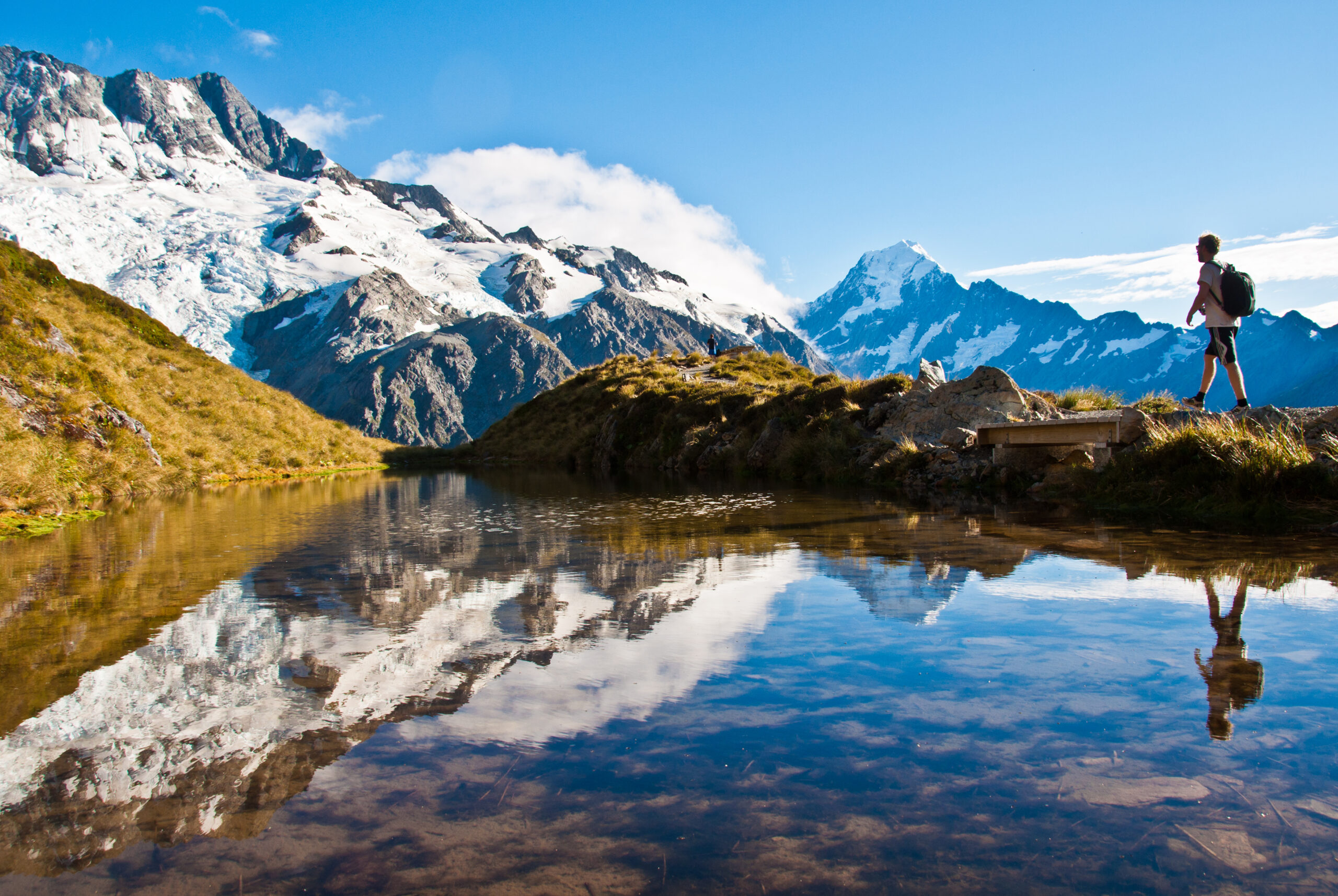 Hiker Overlooking Alpine Lake and Glaciated Mountains A solo hiker with a backpack stands on a grassy ridge overlooking a pristine alpine lake with mirror-like reflections of snow-capped mountains and glaciers in the background, exemplifying scenic hiking and mountaineering destinations.