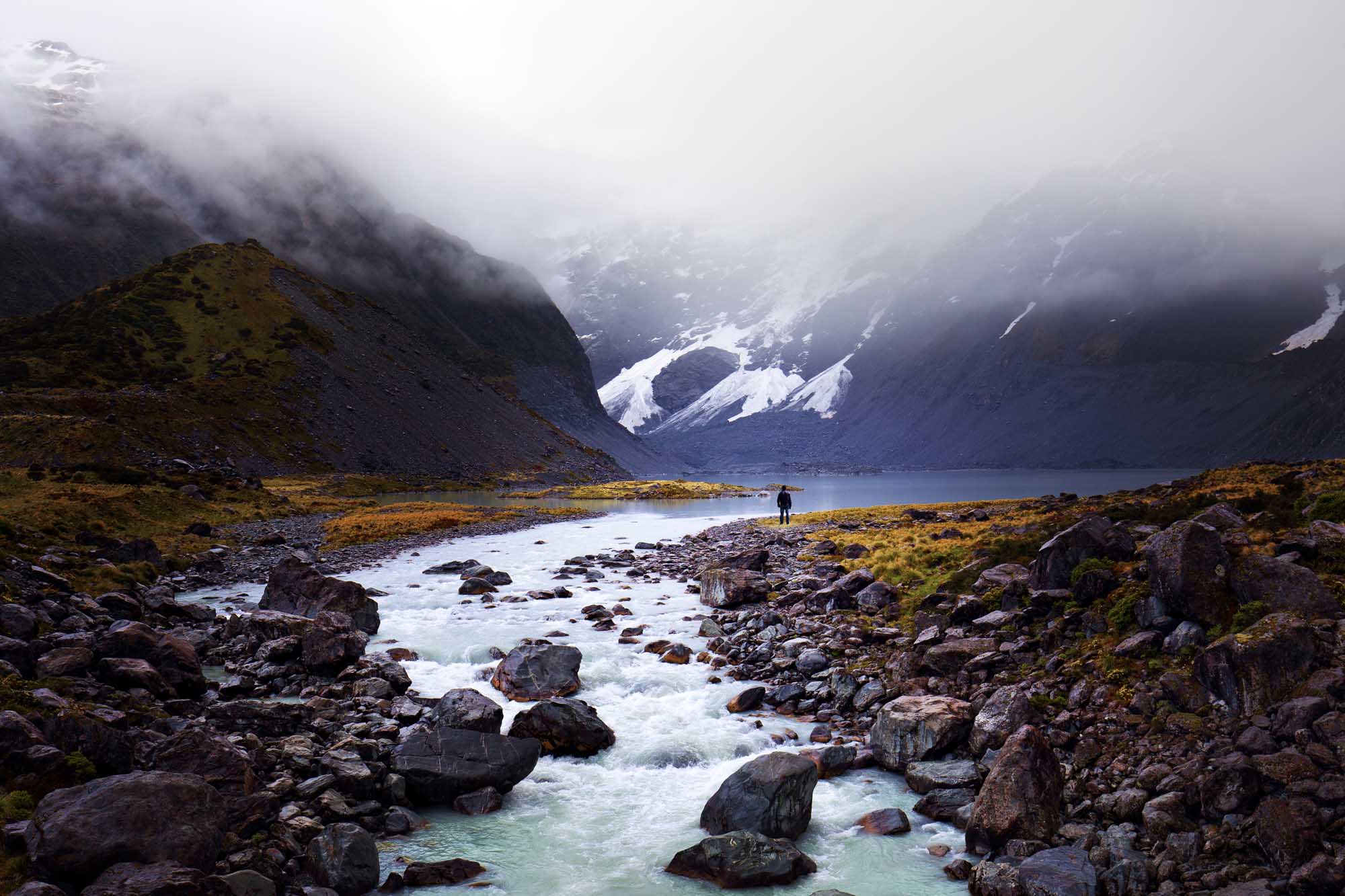 A solitary hiker stands in a remote Arctic valley surrounded by snow-capped mountains shrouded in mist, with a pristine glacial stream flowing across rocky terrain in the foreground. The dramatic landscape features towering peaks with waterfalls and glaciers visible through the fog, creating a stark and otherworldly wilderness scene.