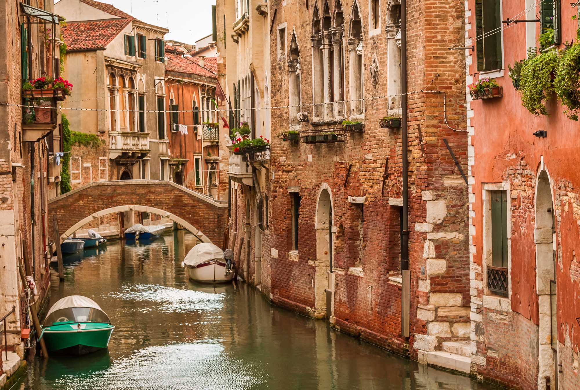 Historic Canal with Boats in Venice A picturesque view of a narrow Venetian canal lined with historic brick and stone buildings, featuring traditional arched bridges, moored boats, and vibrant red facades typical of Venice's Grand Canal architecture.