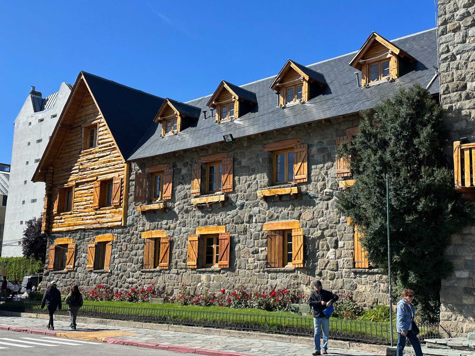 Historic Stone Building with Mansard Roof A striking heritage stone building featuring a mansard roof with dormer windows, golden wooden shutters, and log cabin architectural elements on the left wing. The structure combines stone masonry with timber details and is fronted by manicured gardens with red flowers, attracting visitors on a clear day.