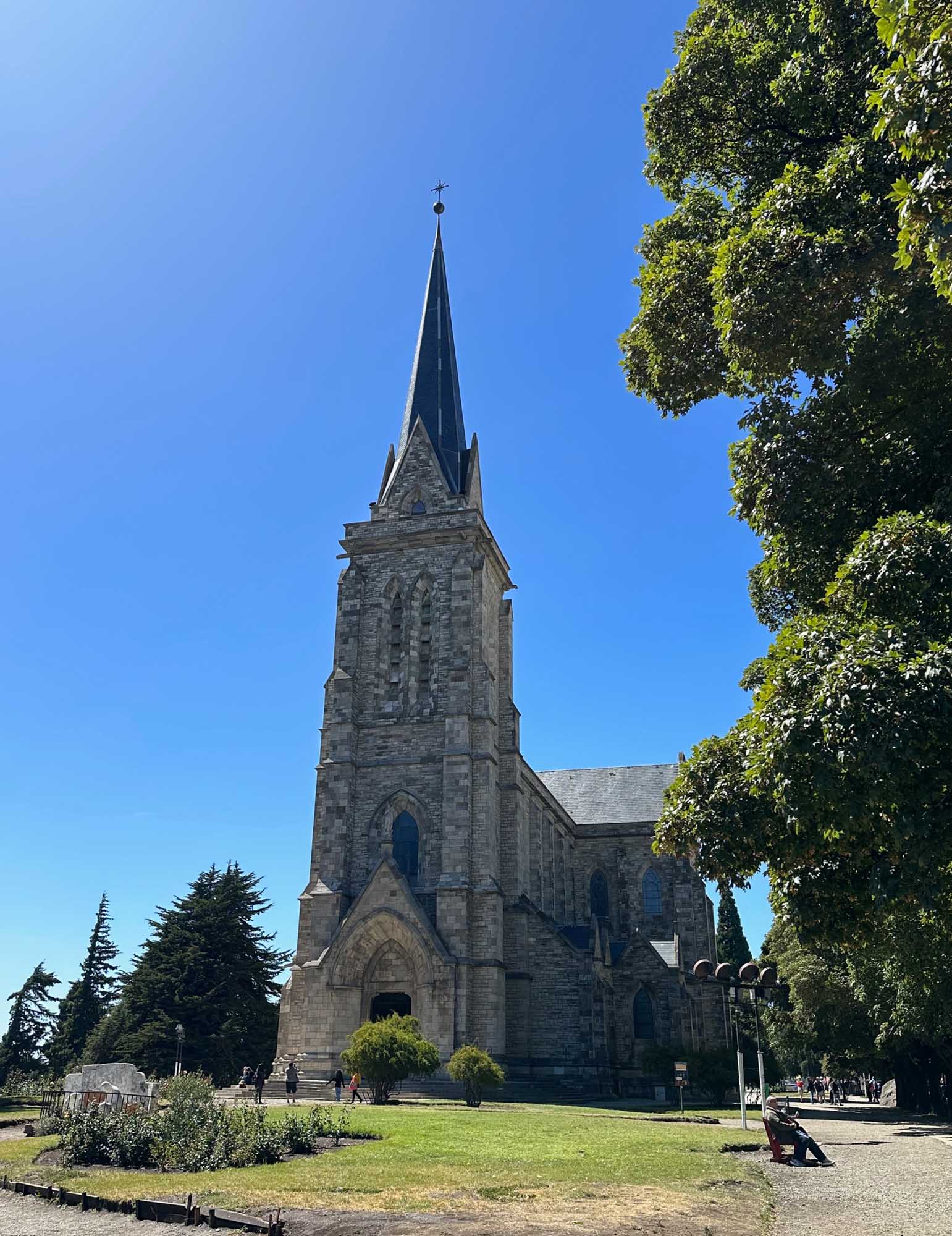Historic Stone Church with Spire in Patagonia A striking Gothic Revival stone church with a tall pointed spire topped by a cross stands prominently against a clear blue sky. The church features ornate stonework, arched windows, and is surrounded by manicured grounds with mature trees and ivy-covered walls, characteristic of early colonial religious architecture in Argentine Patagonia.