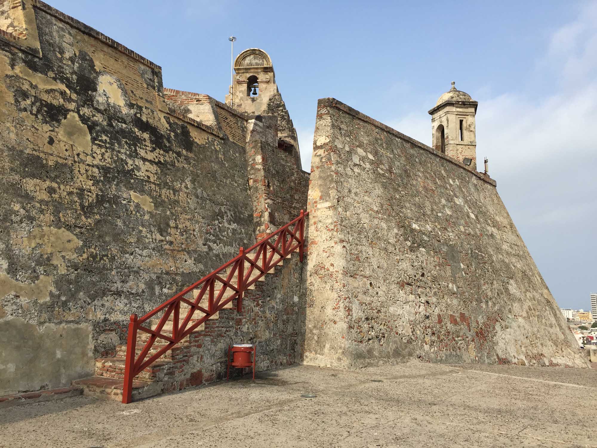 Historic Stone Fort with Bell Tower A weathered fortification with massive stone walls featuring a prominent bell tower and a bright red metal staircase leading up the exterior. The structure shows significant age and patina, typical of colonial-era coastal defense forts.
