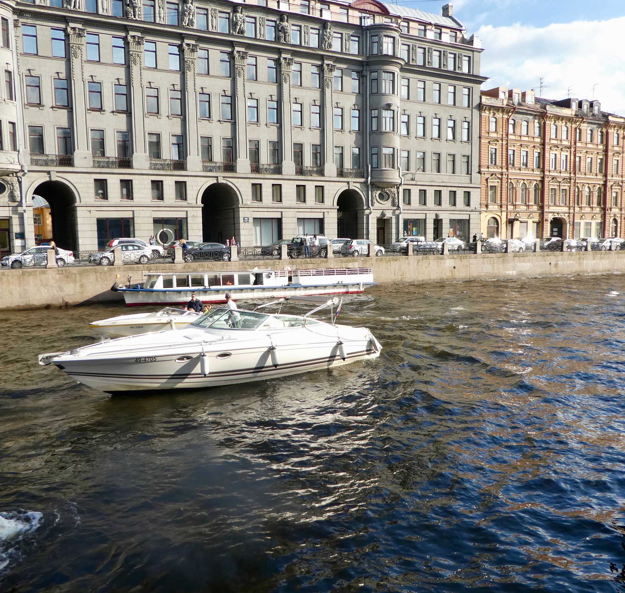Historic Waterfront Buildings and Moored Boats A picturesque waterfront scene featuring elegant historic European architecture with classical stone buildings, ornate columns, and mansard roofs lining the quay. White and blue leisure boats are moored in the calm water, with a passenger tour boat visible along the embankment.
