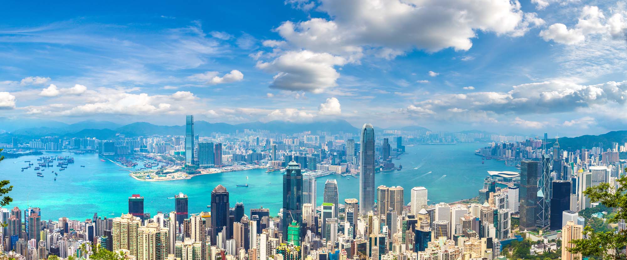A stunning panoramic view of Hong Kong's iconic Victoria Harbour and business district skyline, featuring towering skyscrapers, turquoise blue waters dotted with vessels, and lush green mountains in the background under a beautiful blue sky with white clouds.