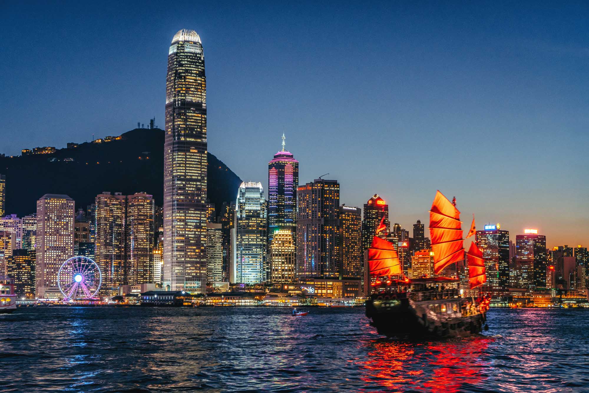 Hong Kong Skyline with Traditional Junk Boat at Dusk A stunning twilight view of Hong Kong's illuminated skyline featuring iconic skyscrapers including the International Finance Centre, with a traditional red-sailed junk boat prominently positioned in Victoria Harbour. The scene captures the contrast between modern architecture and historic maritime culture, with the Star Ferry Wheel and cityscape lights reflected in the water.
