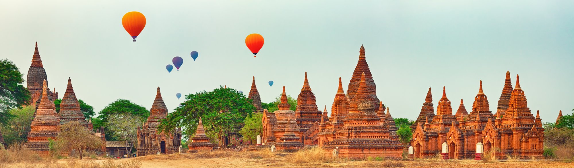 Hot Air Balloons Over Ancient Temples A stunning panoramic view of multiple ancient Buddhist temples with distinctive brick spires dotting a landscape, with colorful hot air balloons floating gracefully in the sky above. The scene captures the magical atmosphere of a popular Southeast Asian destination during sunrise or early morning.