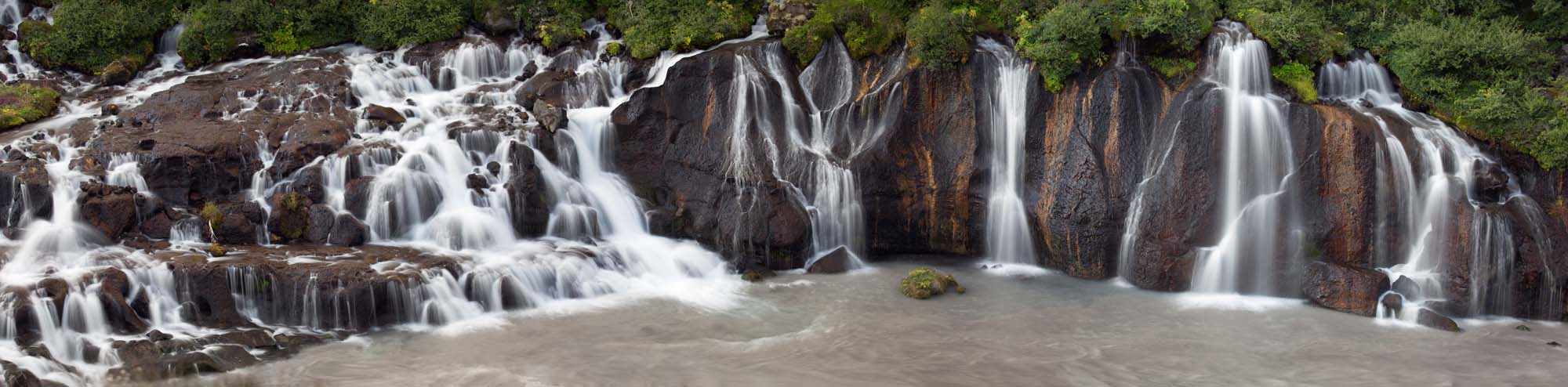 A stunning cascade of multiple waterfalls flowing over dark basalt rock formations into a milky glacial river pool, surrounded by lush green vegetation. The image captures the dramatic natural beauty of Iceland's Hraunfossar waterfall system with its distinctive tiered drops and powerful water flow.