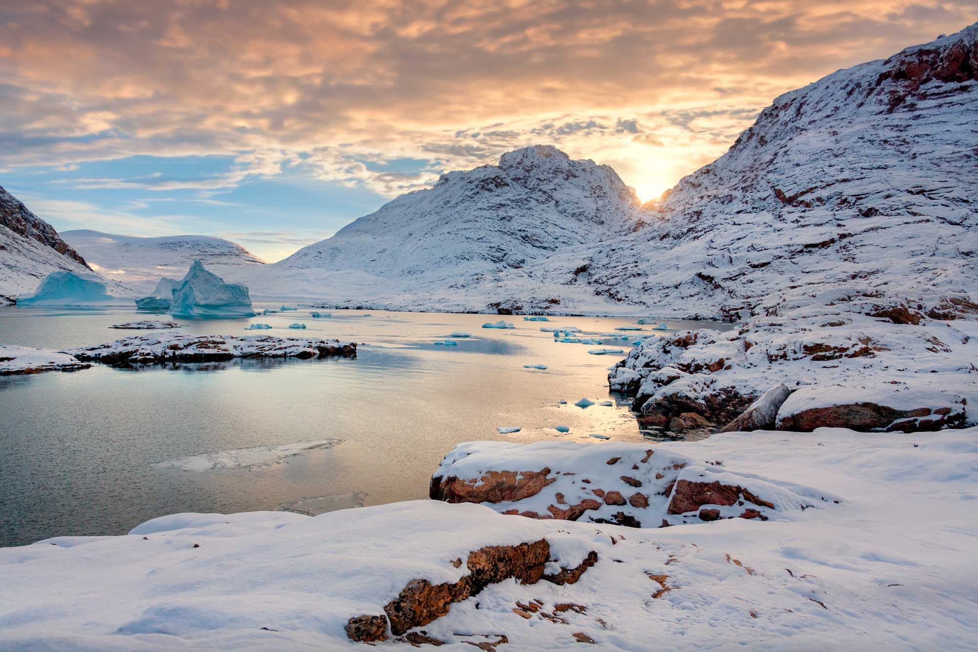 Hurry Inlet at Dusk, East Greenland A stunning Arctic landscape featuring snow-covered mountains framing a frozen inlet dotted with turquoise icebergs at sunset. The scene captures the dramatic beauty of King Christian X Land on Greenland's east coast, with golden light illuminating the peak between towering mountain slopes.