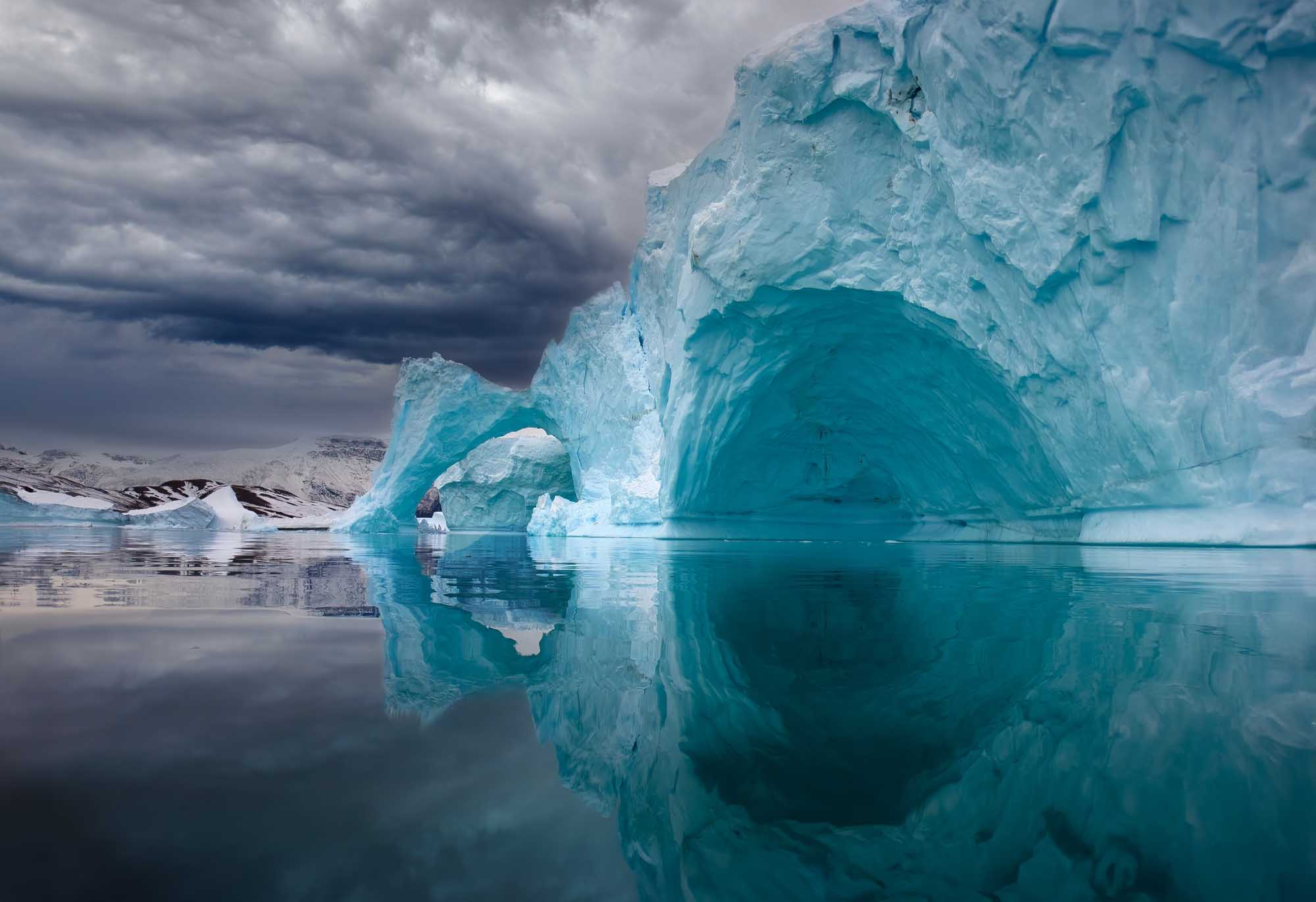 Iceberg with Arch Reflection in East Greenland A striking turquoise iceberg featuring a natural arch formation floats in calm, mirror-like waters off the coast of East Greenland. Snow-capped mountains are visible in the background beneath a dramatic cloudy sky, creating a serene Arctic landscape.