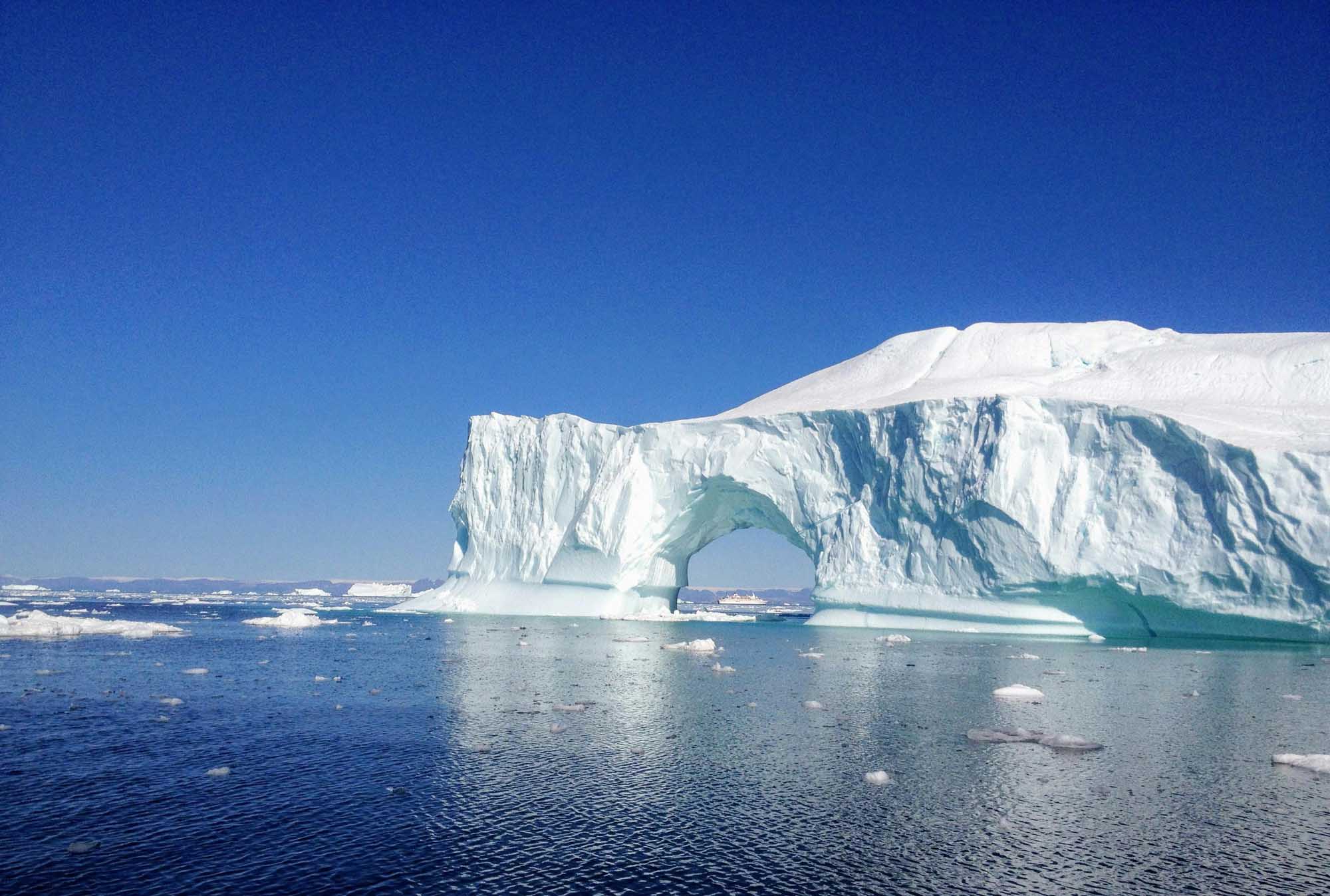Iceberg with Natural Arch in Arctic Waters A massive tabular iceberg featuring a striking natural arch formation floats in calm Arctic waters under a clear blue sky. The ice structure displays brilliant white and turquoise hues, with smaller ice chunks scattered across the surrounding water, characteristic of polar regions during the melting season.