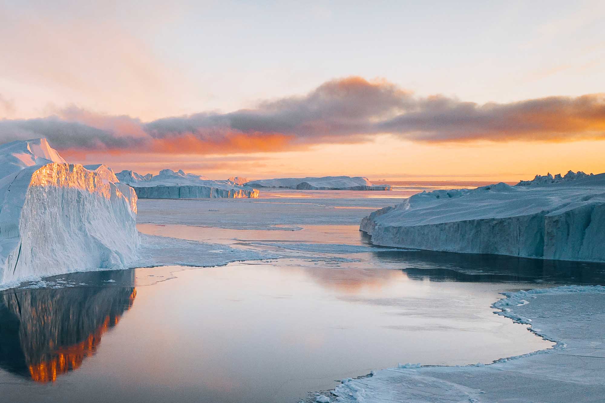 Icebergs and Frozen Sea at Arctic Sunrise A stunning arctic landscape featuring massive icebergs with white and blue hues reflected in the partially frozen sea during golden sunrise. Dramatic cloudy sky and distant ice formations create a pristine, remote polar scenery characteristic of Greenland's coastal regions.