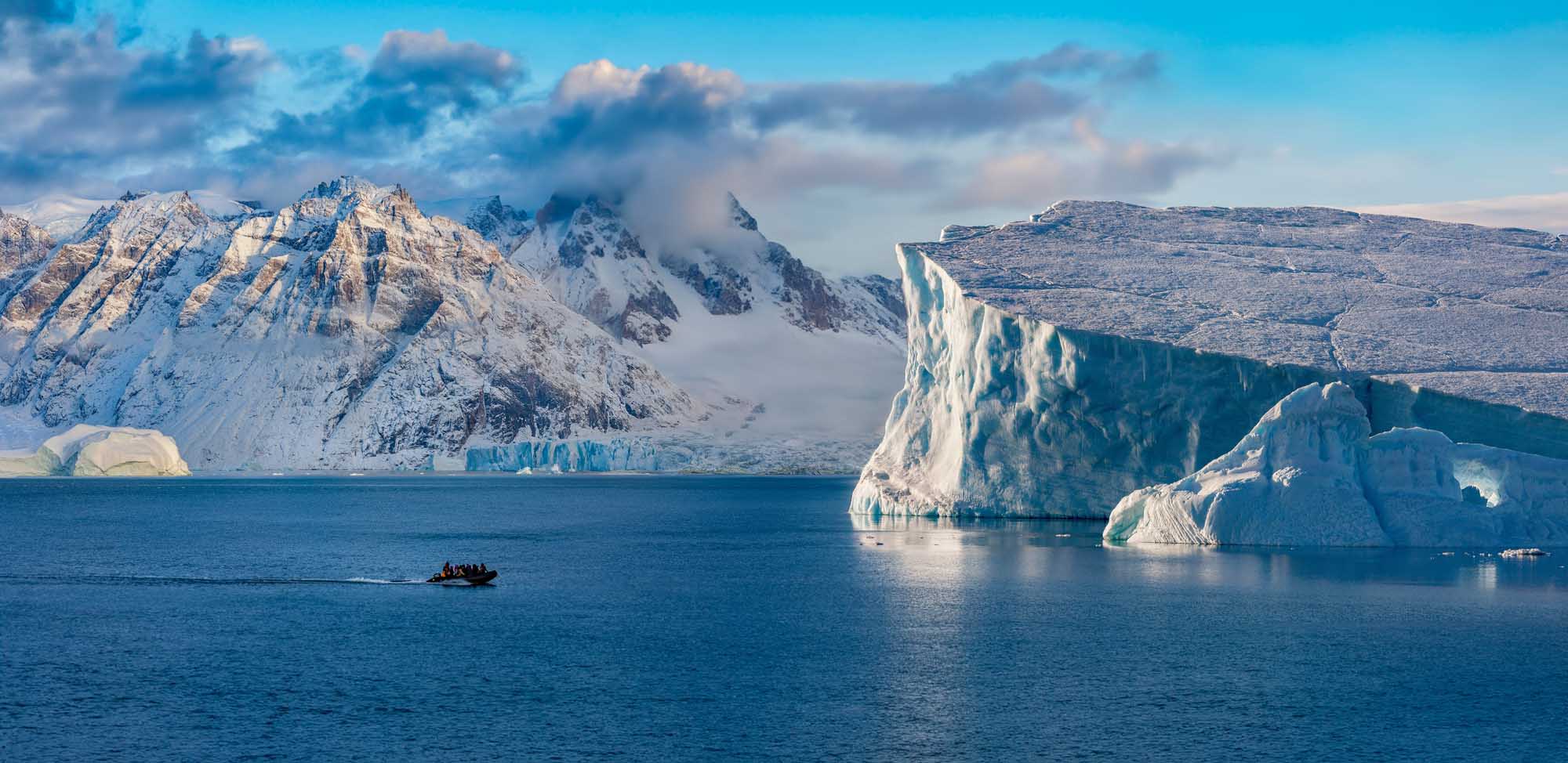 Icebergs and Mountains in Scoresbysund, Greenland A breathtaking Arctic landscape featuring towering icebergs with striking turquoise hues floating in deep blue waters, snow-capped mountains in the background, and a small inflatable boat with adventure tourists exploring the pristine fjord during golden afternoon light.