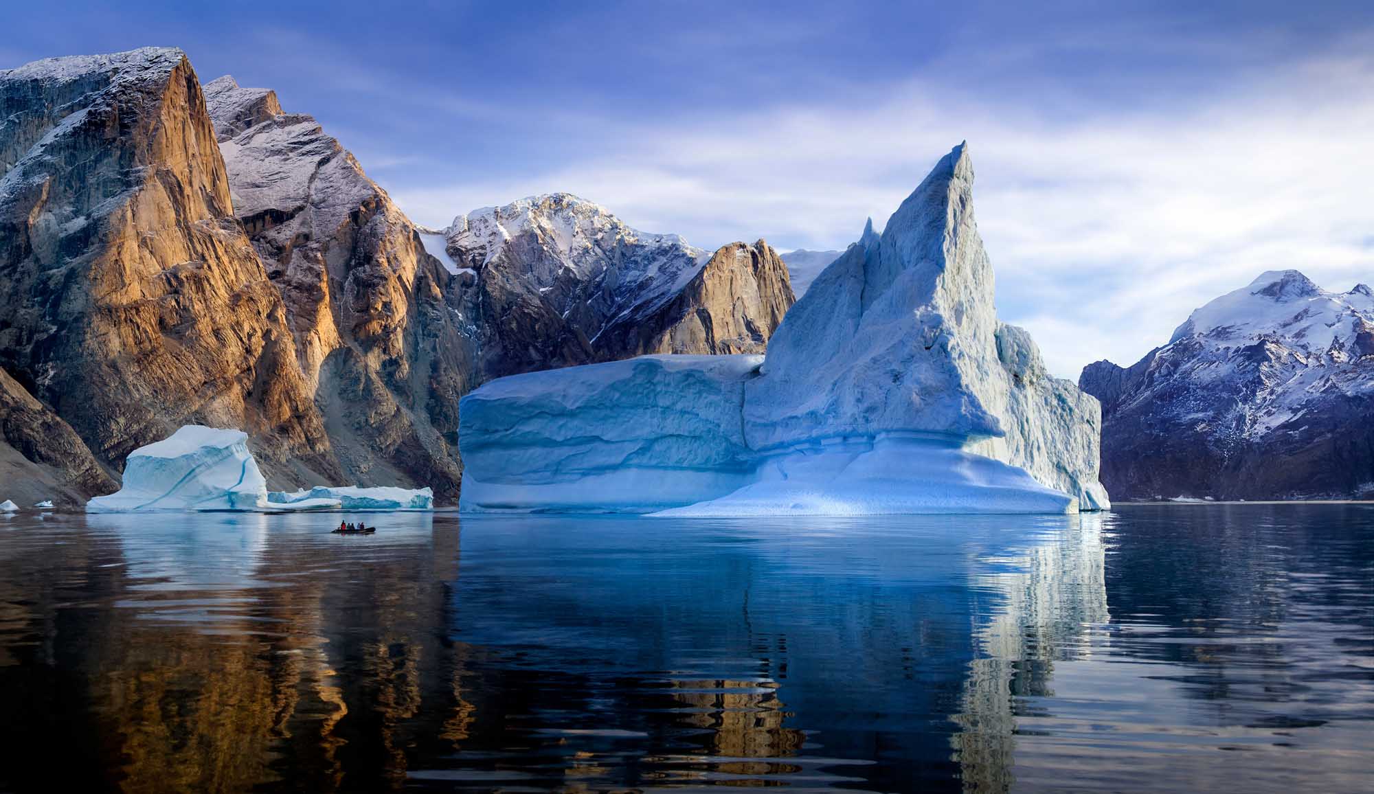 Icebergs in Franz Joseph Fjord, Greenland A stunning Arctic landscape featuring towering icebergs with dramatic blue and white coloring floating in calm fjord waters, surrounded by snow-capped mountains. A small boat with adventure tourists can be seen exploring among the massive ice formations in eastern Greenland.
