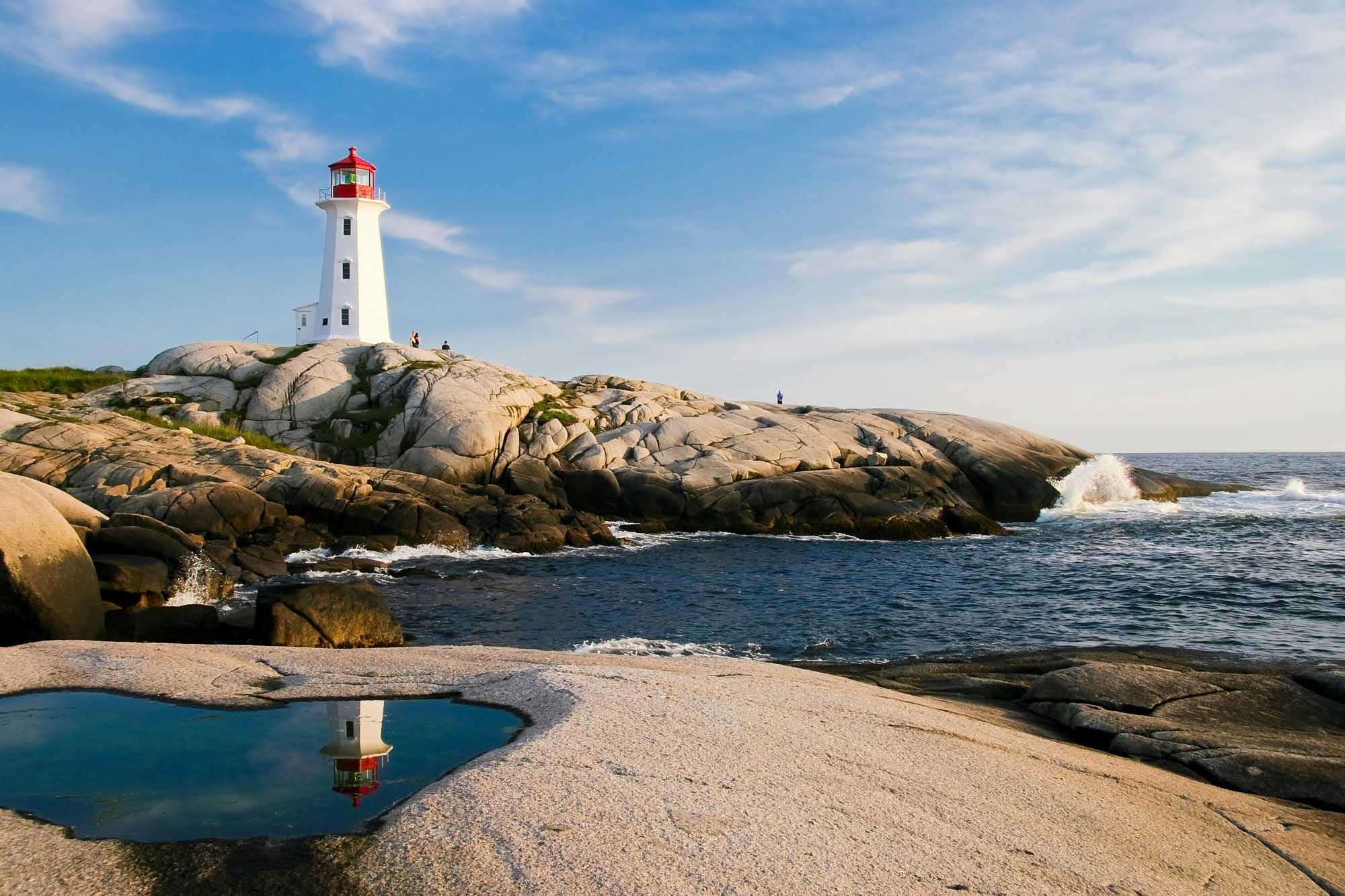 A classic white lighthouse with a red lantern room stands prominently on a rugged granite rocky outcrop overlooking the Atlantic Ocean. The scene captures a serene coastal landscape with a natural tidal pool reflecting the lighthouse, scattered boulders, crashing waves, and a clear blue sky with wispy clouds.