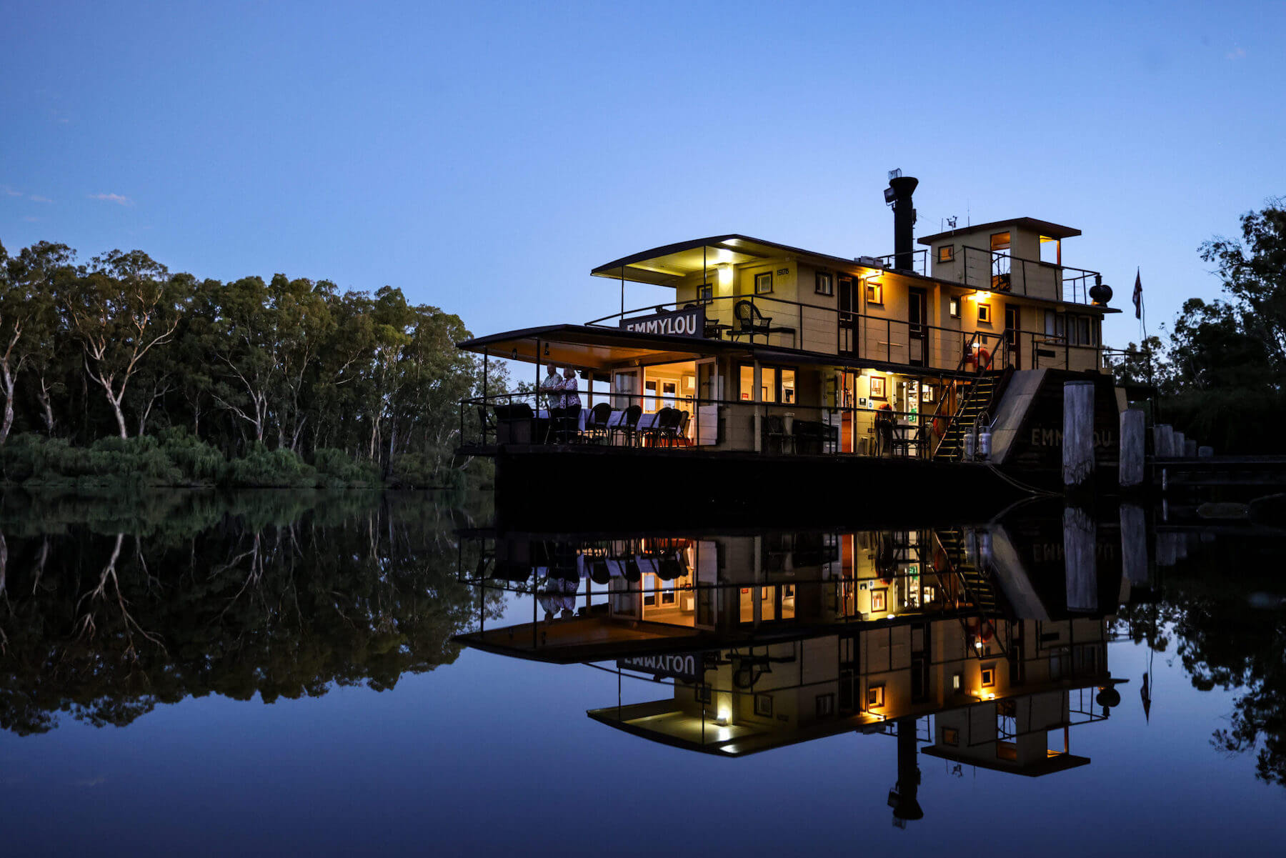 Illuminated Riverboat at Twilight with Reflections A striking nighttime image of the PS Emmylou, a heritage riverboat, beautifully lit against a twilight sky and mirrored perfectly in the still water below. The vessel is moored on a peaceful river lined with eucalyptus trees, creating a serene and atmospheric scene of river cruise tourism.