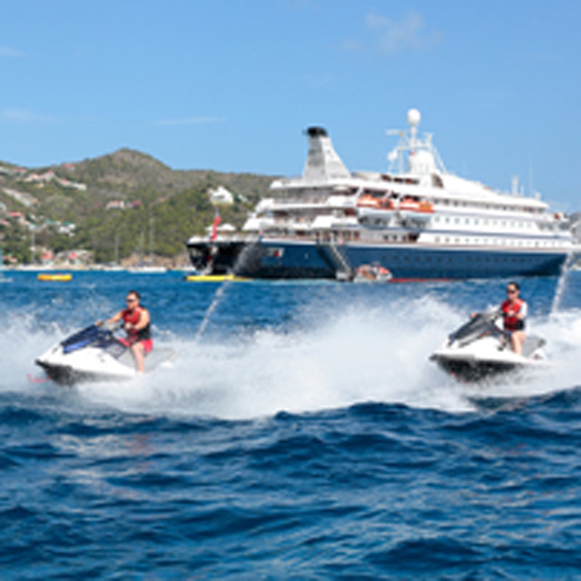 Jet Ski Adventure Near Cruise Ship Two jet skiers in red life vests enjoy water sports in front of a large cruise ship anchored in a tropical bay, with a forested hillside visible in the background under clear blue skies.