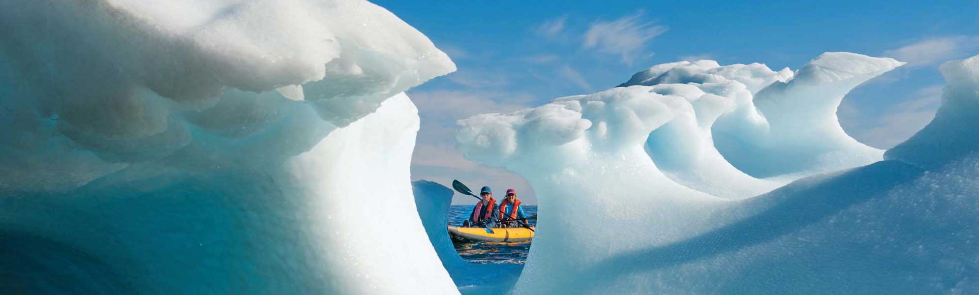 Two adventurers in a yellow kayak navigate through towering turquoise and white icebergs in Arctic waters under clear blue skies, showcasing the dramatic scale and beauty of polar ice formations.
