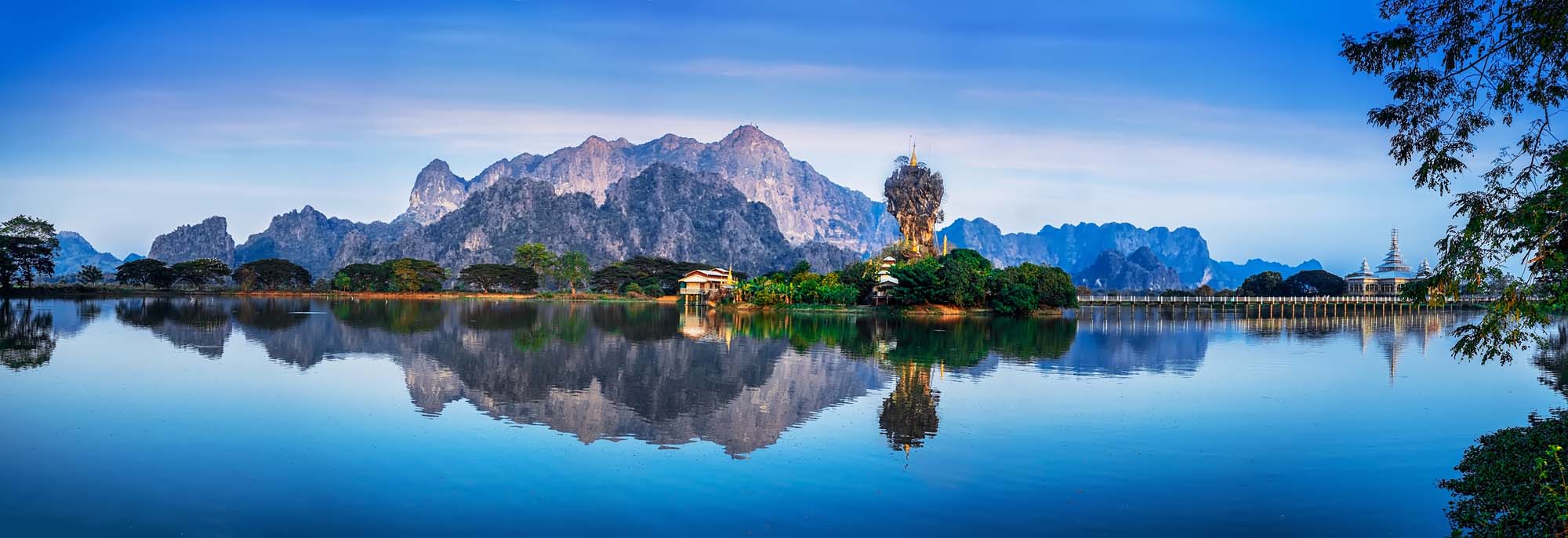 Kyauk Kalap Pagoda Reflected in Still Water A stunning panoramic view of the iconic Kyauk Kalap Pagoda perched atop a dramatic limestone rock formation in Hpa-An, Myanmar, perfectly mirrored in the calm waters below. The golden pagoda and surrounding karst mountains create a serene and picturesque landscape under clear blue skies.