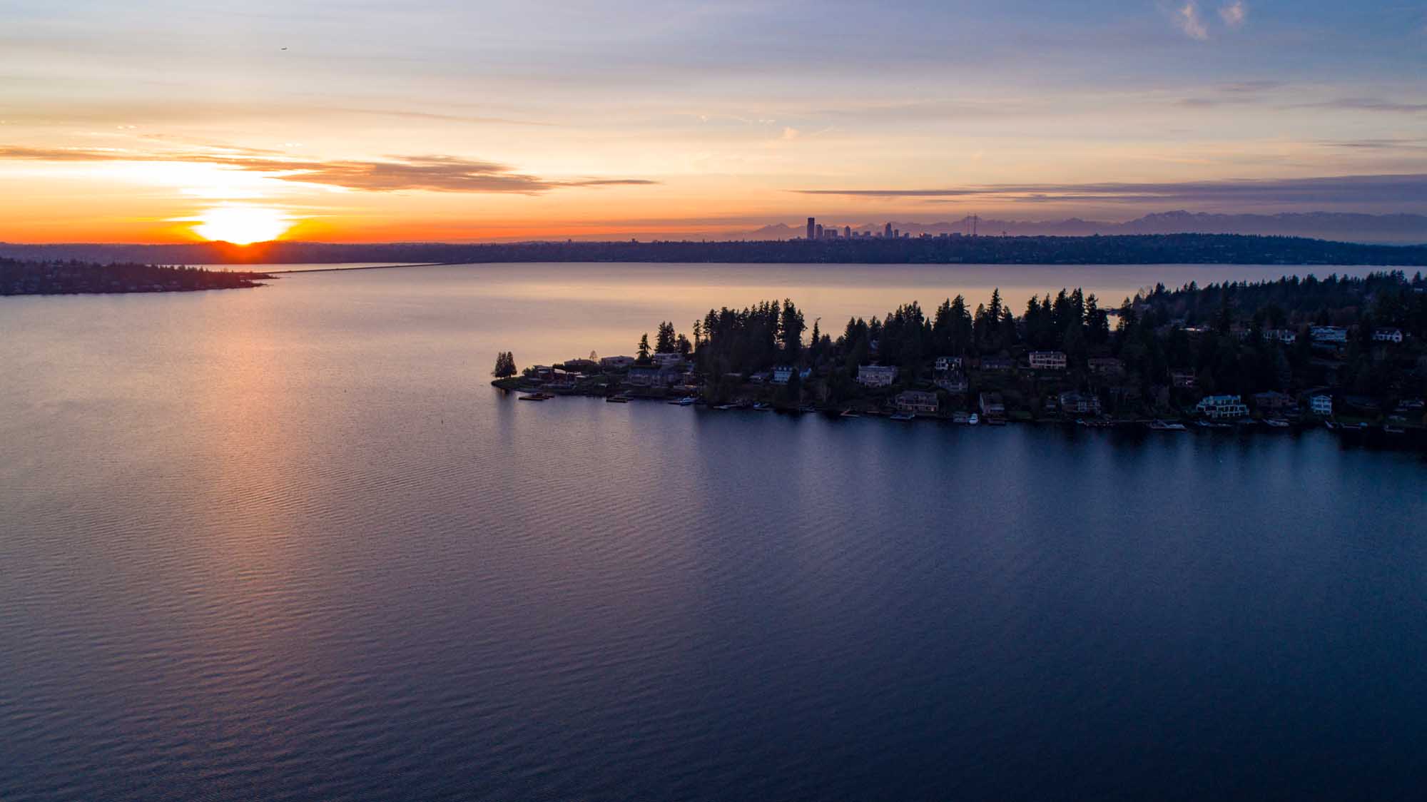 A breathtaking sunset view over Lake Washington, featuring a residential waterfront community with dense evergreen forests in the foreground and the Seattle skyline silhouetted against a golden and purple sky across the water. This scenic vista captures the natural beauty of the Pacific Northwest with calm water reflections and the iconic Puget Sound region landscape.
