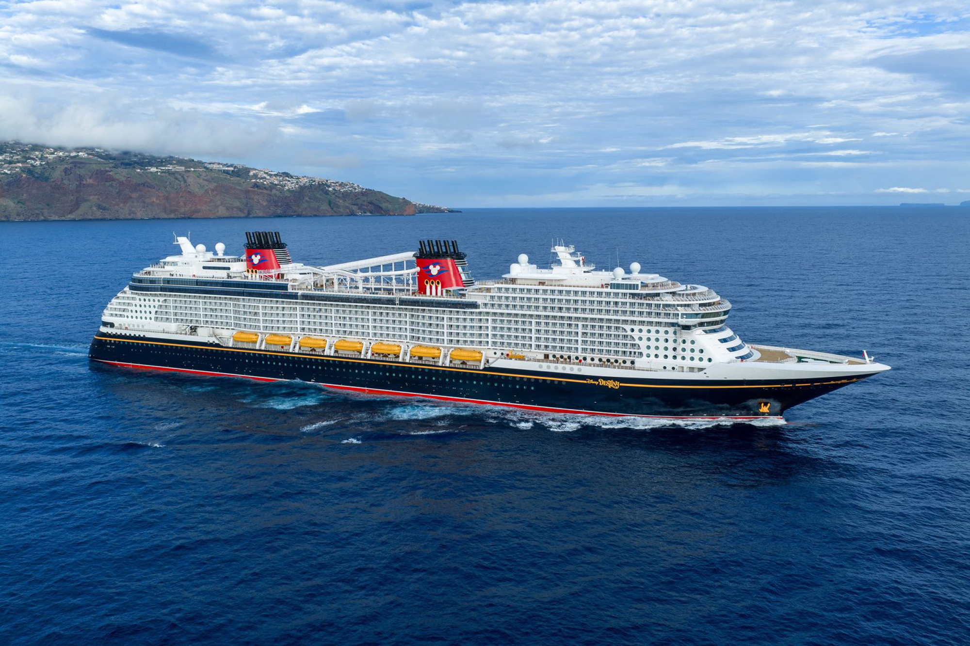 A massive modern cruise ship with distinctive red funnels featuring Mickey Mouse branding is anchored in brilliant blue waters off a volcanic island with lush settlements on its slopes. The vessel features a black hull with red striping, white superstructure, and yellow lifeboats, photographed under partly cloudy skies with a Mediterranean or Atlantic island landscape in the background.